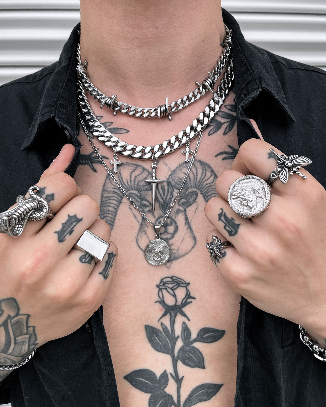 Wearing the Protect Ya Neck Stack, a tattooed individual with layered silver necklaces and rings—featuring animals, crosses, and a bold fallen angel medallion—holds open their shirt to reveal a ram’s head chest tattoo and rose hand ink.