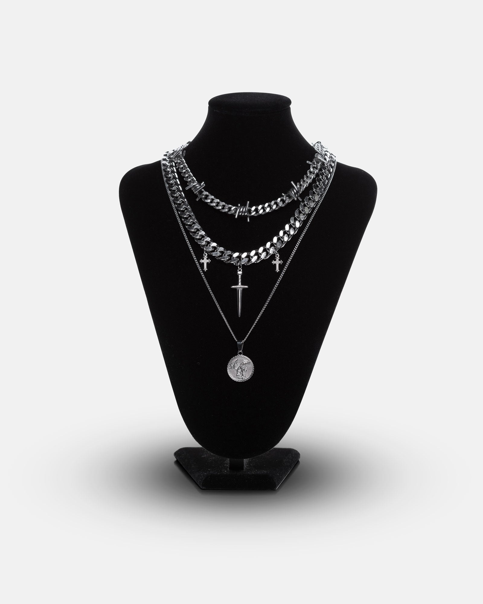 The Protect Ya Neck Stack is displayed on a black velvet bust, featuring layered silver necklaces—a barbed wire Cuban chain, small cross pendants, and a fallen angel medallion—set against a plain white background.