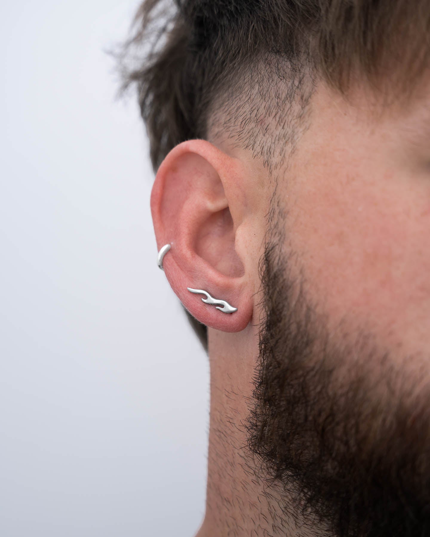 A close-up shows a person with short brown hair and a trimmed beard wearing "Pyro" Flame Earrings, made from hypoallergenic stainless steel, and a silver wavy ear cuff. The background is plain and light-colored.
