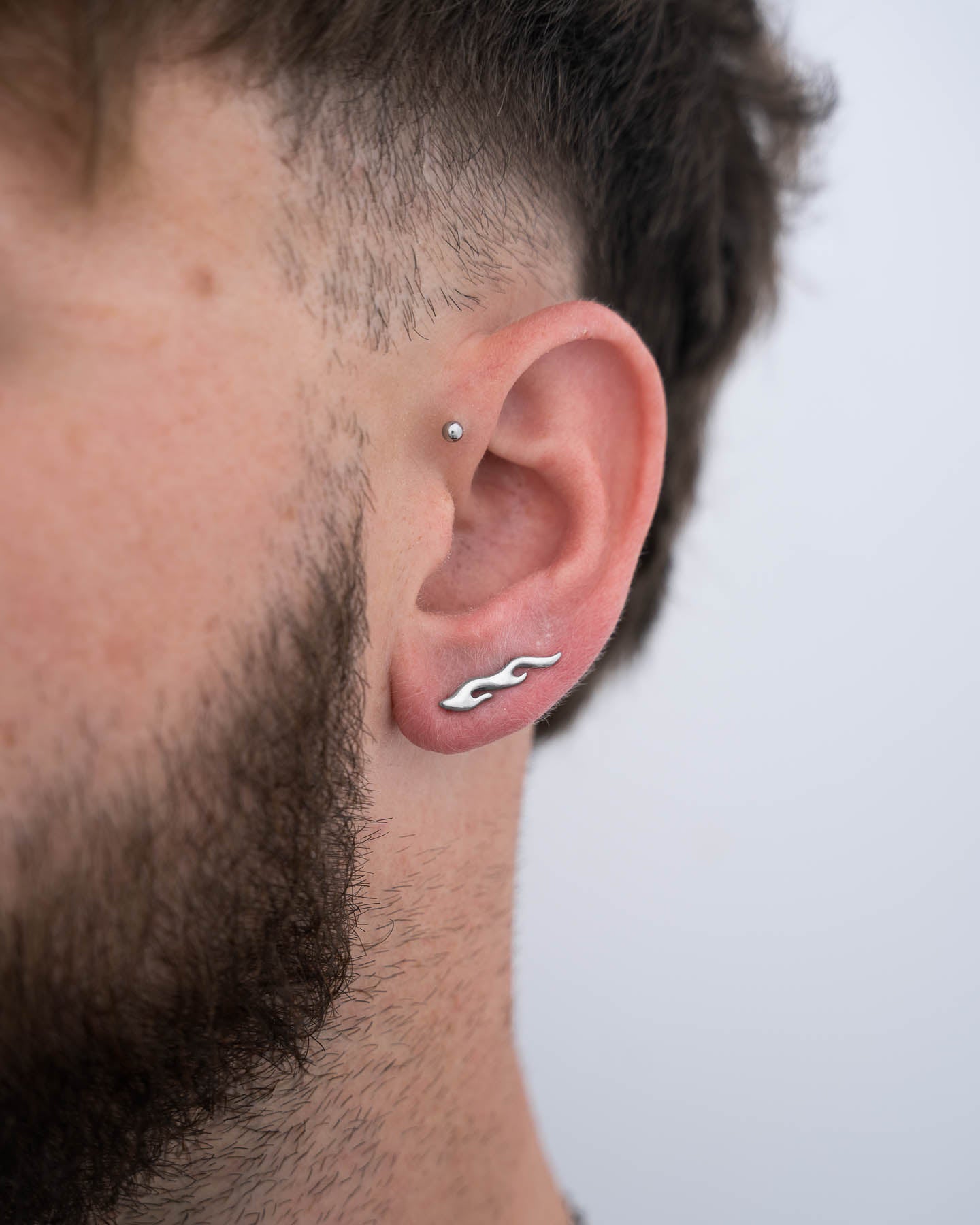 Close-up of a person with short brown hair and a trimmed beard wearing hypoallergenic stainless steel earrings: a small silver stud in the cartilage and a "Pyro" Flame Earring in the lobe.