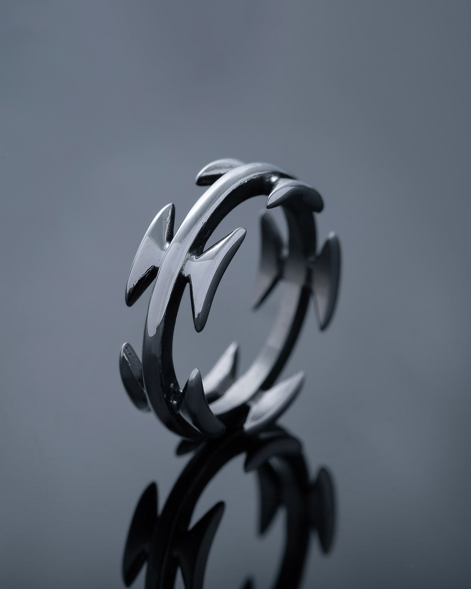The Razor Wire Ring features pointed arrow-shaped spikes evenly spaced around a stainless steel band, photographed on a reflective dark surface—a bold piece of streetwear jewelry.