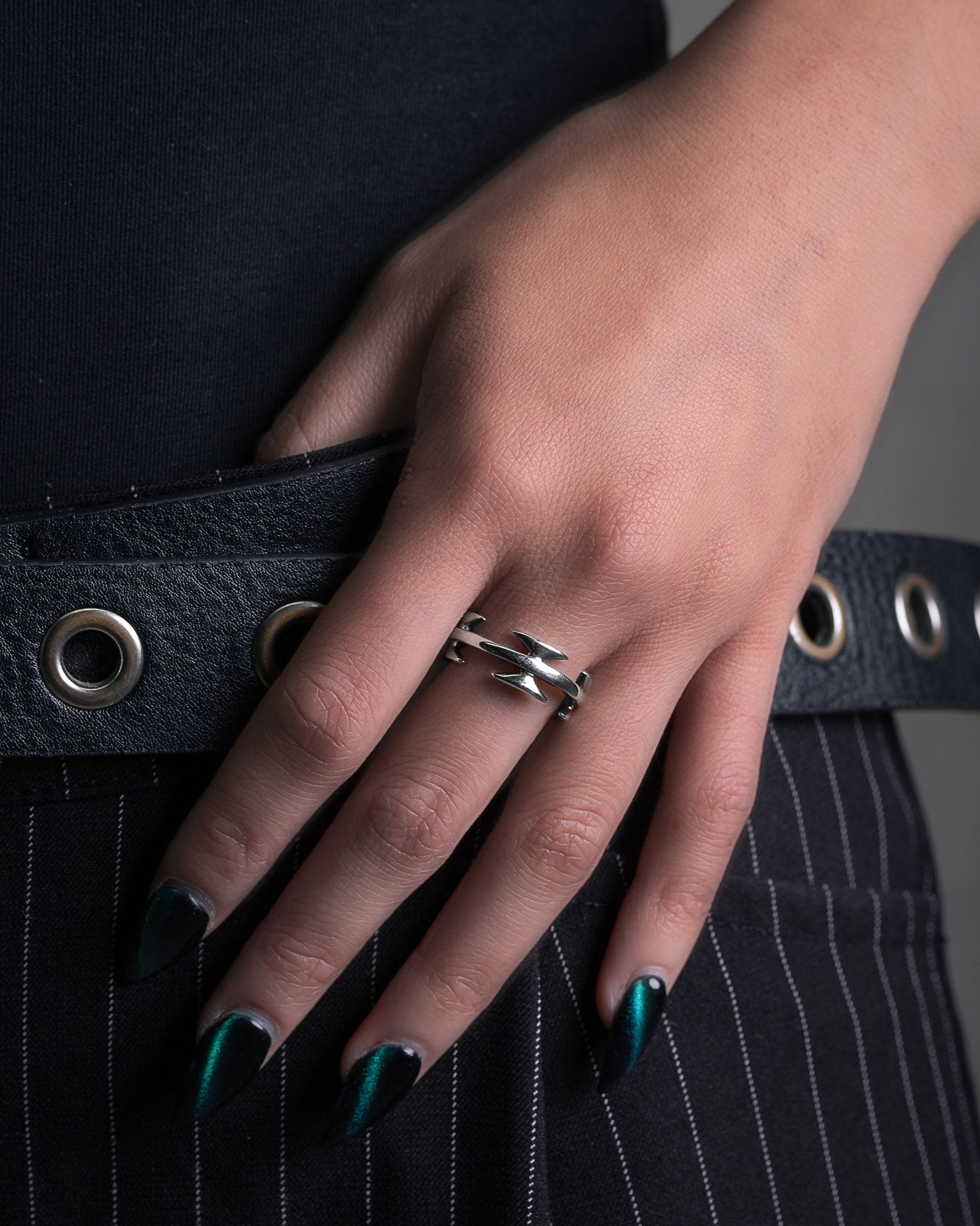 A hand with dark green, pointed nails rests on a hip, showcasing the Razor Wire Ring—an edgy streetwear accessory. The look is finished with pinstripe pants and a black belt with silver eyelets.