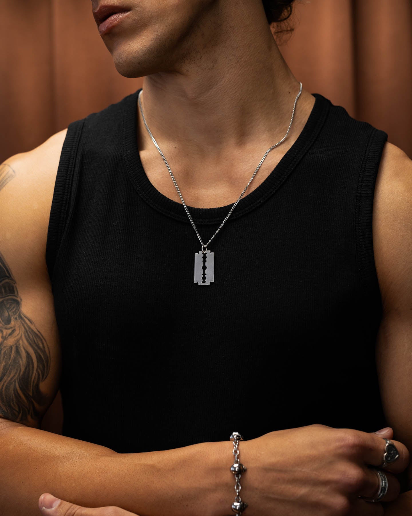 Wearing a black sleeveless shirt, punk bracelet, and a Razor Blade Pendant stainless steel necklace, the tattooed person stands against a brown background.