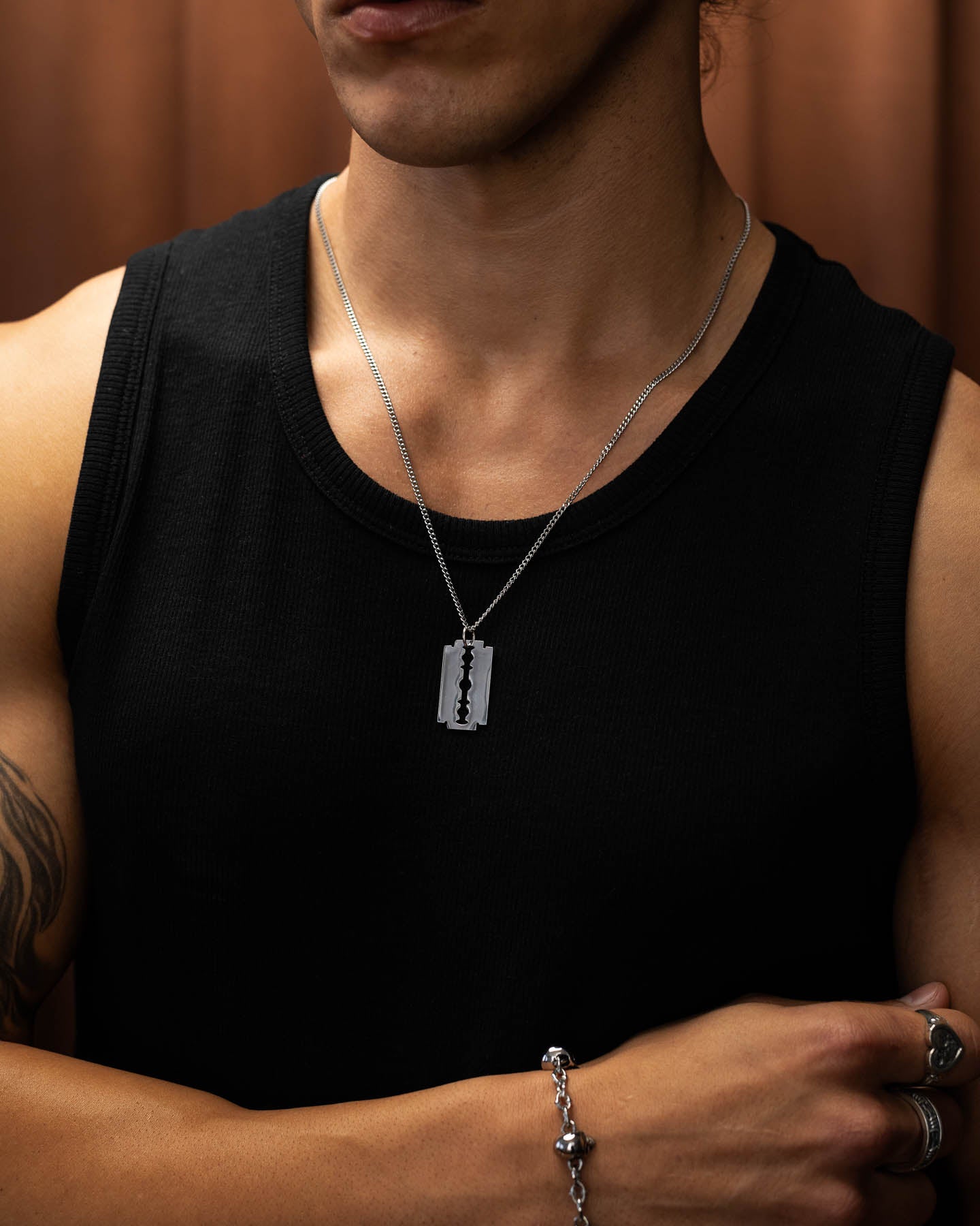 A person wearing a sleeveless black shirt, silver bracelet, and the Razor Blade Pendant stands with one arm crossed. The softly blurred, warm-toned background highlights their edgy punk jewelry look.