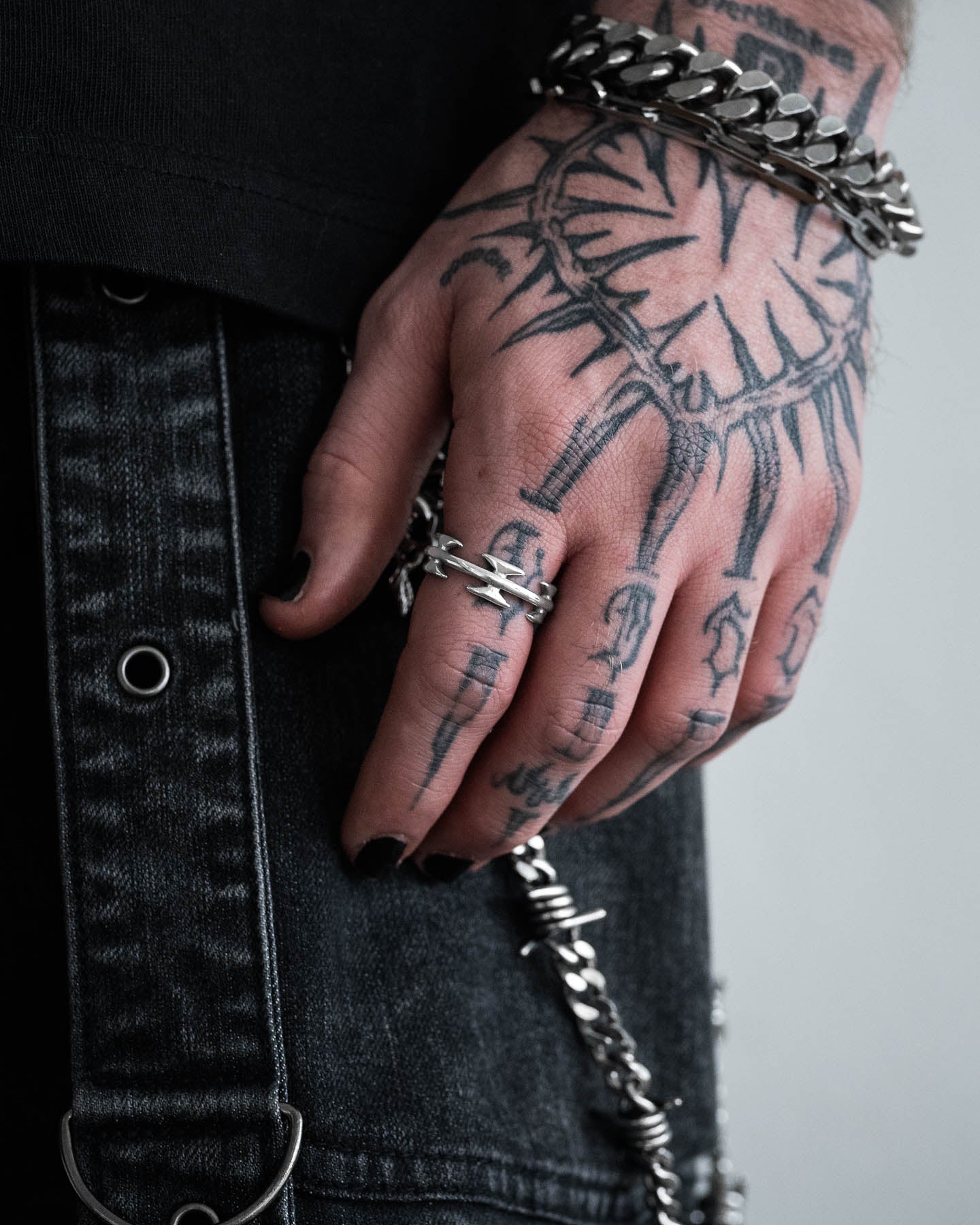 A close-up of a hand with black nail polish, silver rings including the Razor Wire Ring, bracelets, and intricate black tattoos. Black denim with metal details, chains, and edgy streetwear jewellery complete the look.
