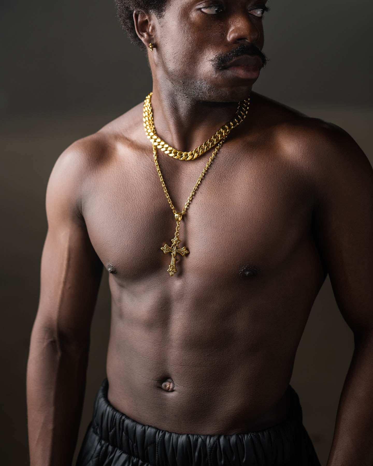 A shirtless man with dark skin wears black pants and two gold chains, including "The Ruins" Pendant (Gold), as he stands against a dark background, looking to his left.