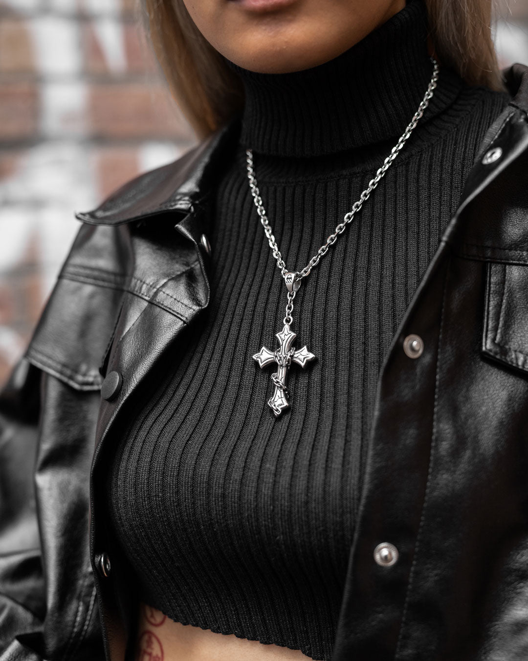 Wearing a black ribbed turtleneck crop top and black leather jacket, the person accessorizes with "The Ruins" Pendant. The background has blurred brick.