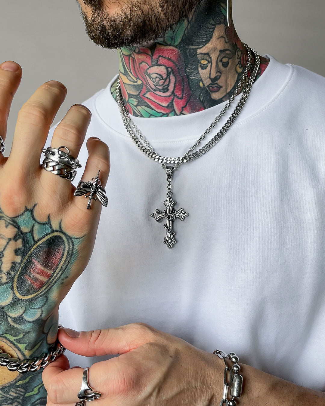 A person in a white shirt showcases tattooed hands and neck, wearing multiple silver rings and "The Ruins" 6mm Set—stainless steel Cuban link chains, one with a detailed gothic cross pendant.