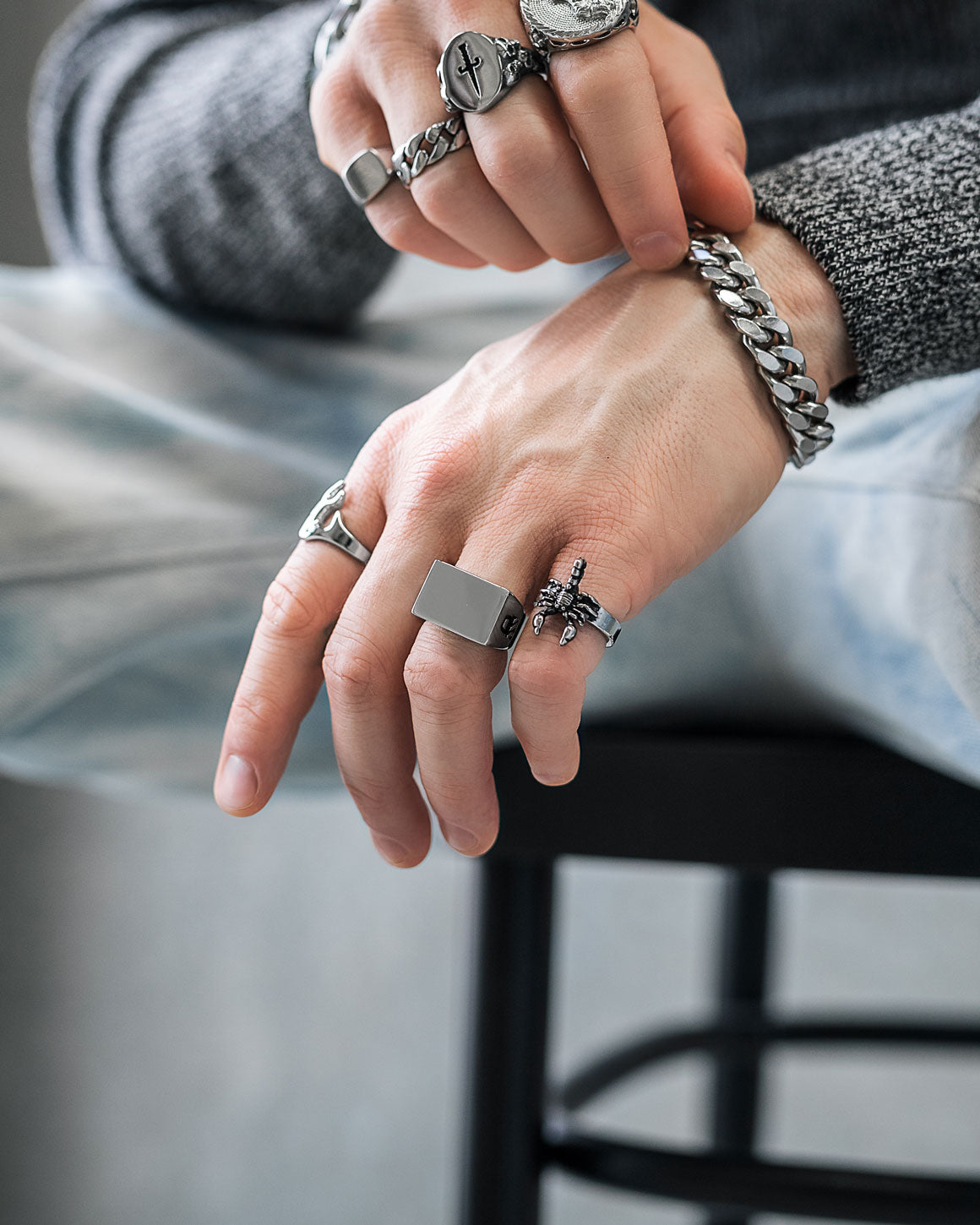 A close-up of hands wearing multiple silver rings, including the Scorpion Ring, and a chunky chain bracelet, resting casually on their lap while seated on a black stool.