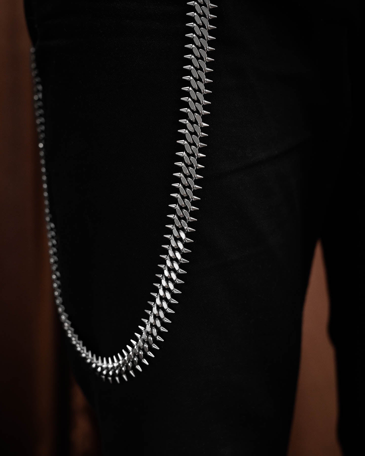 A close-up of the Statement Collective "Screwed Up" 9mm Spiked Cuban Wallet Chain, crafted from 316L stainless steel, attached to black pants and hanging in a curve against a dark background.