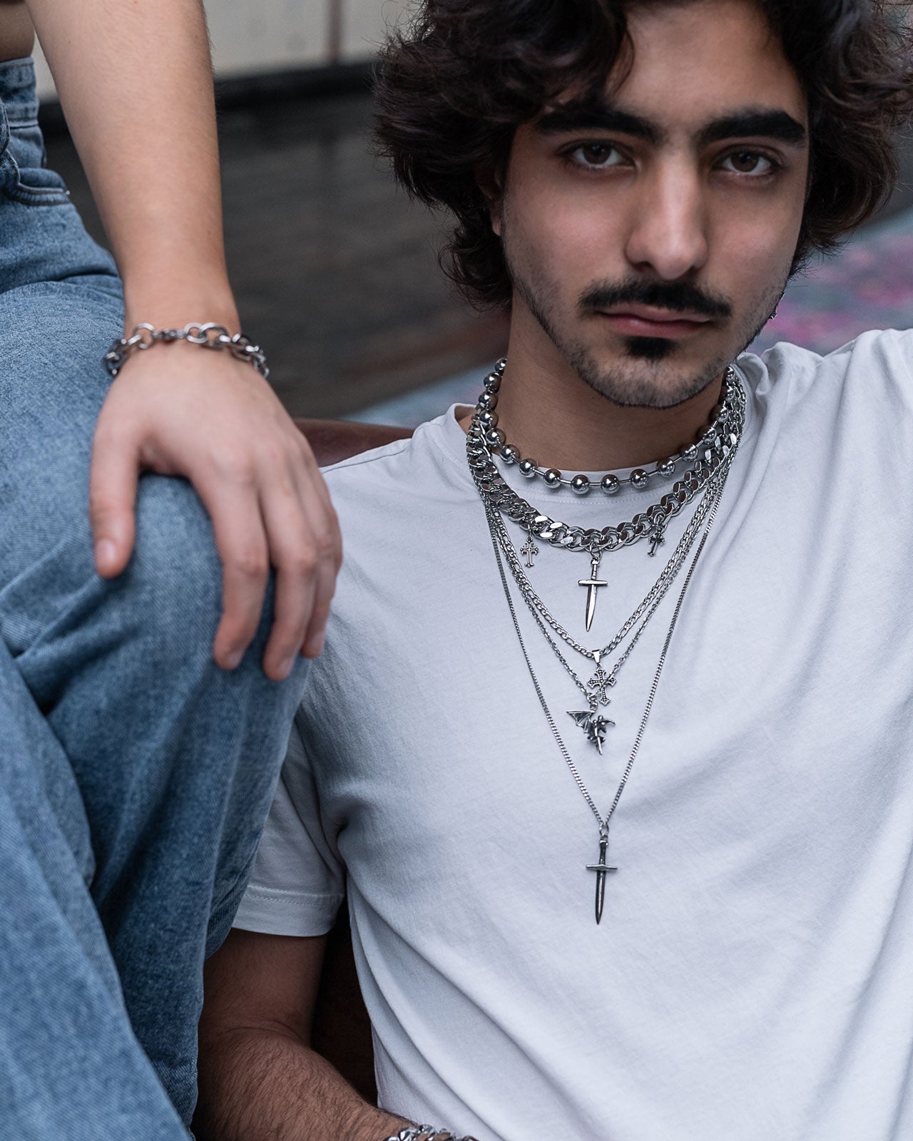A young man with dark hair and a mustache wears a white t-shirt and the Sharp Objects Stack—a set of striking silver necklaces with bold pendants. Next to him, someone in jeans rests an arm on his shoulder; both showcase standout silver jewelry.