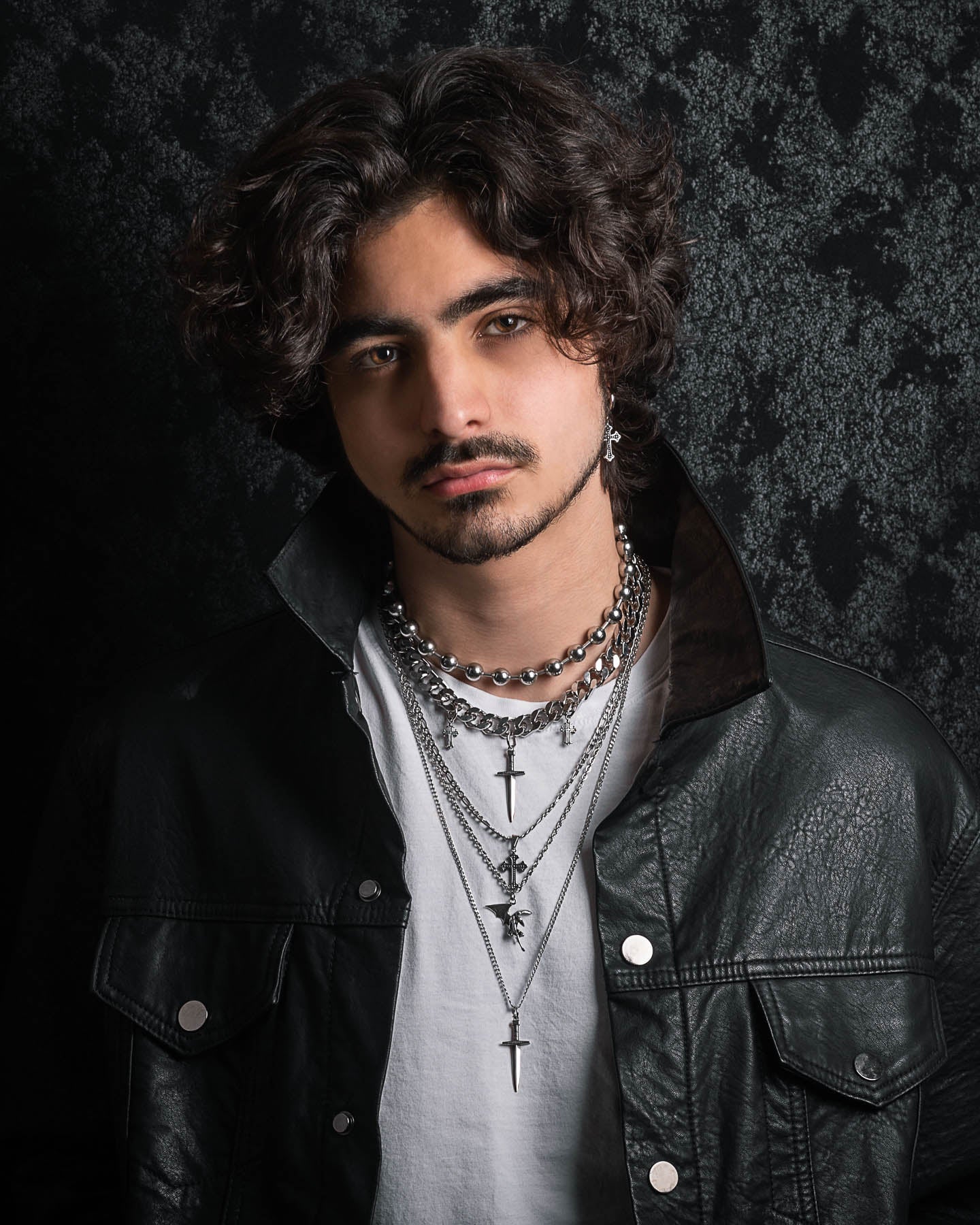A young man with wavy dark hair and a mustache wears the Sharp Objects Stack—a set of layered silver chains featuring crosses and a bold statement necklace—over a white shirt and black leather jacket against a dark, textured background.