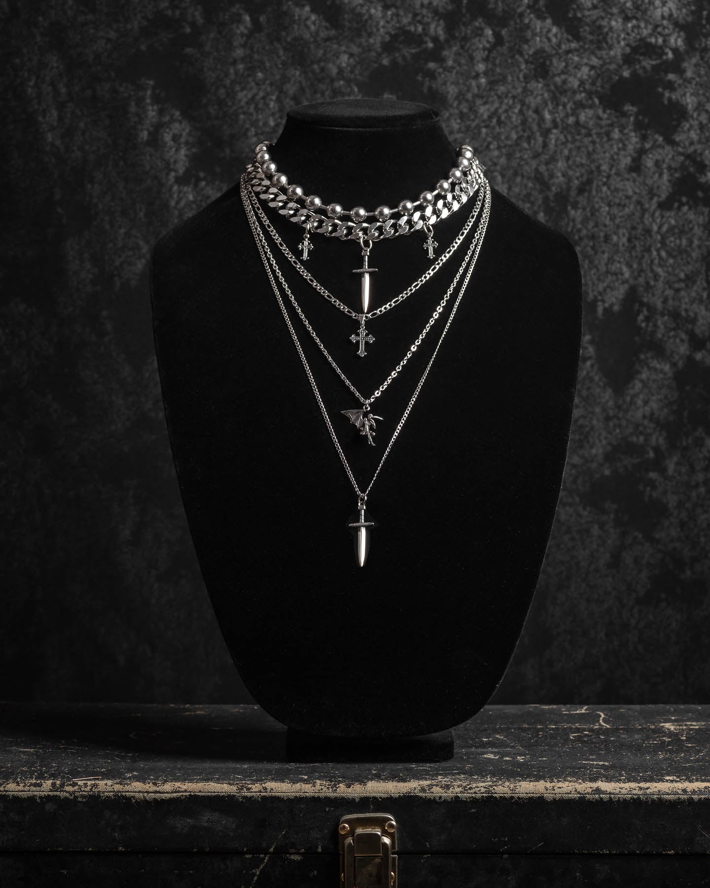 The Sharp Objects Stack features four layered silver pendant necklaces—crosses, spikes, and a statement Cuban link chain—displayed on a black velvet bust atop a vintage case against a dark textured background.