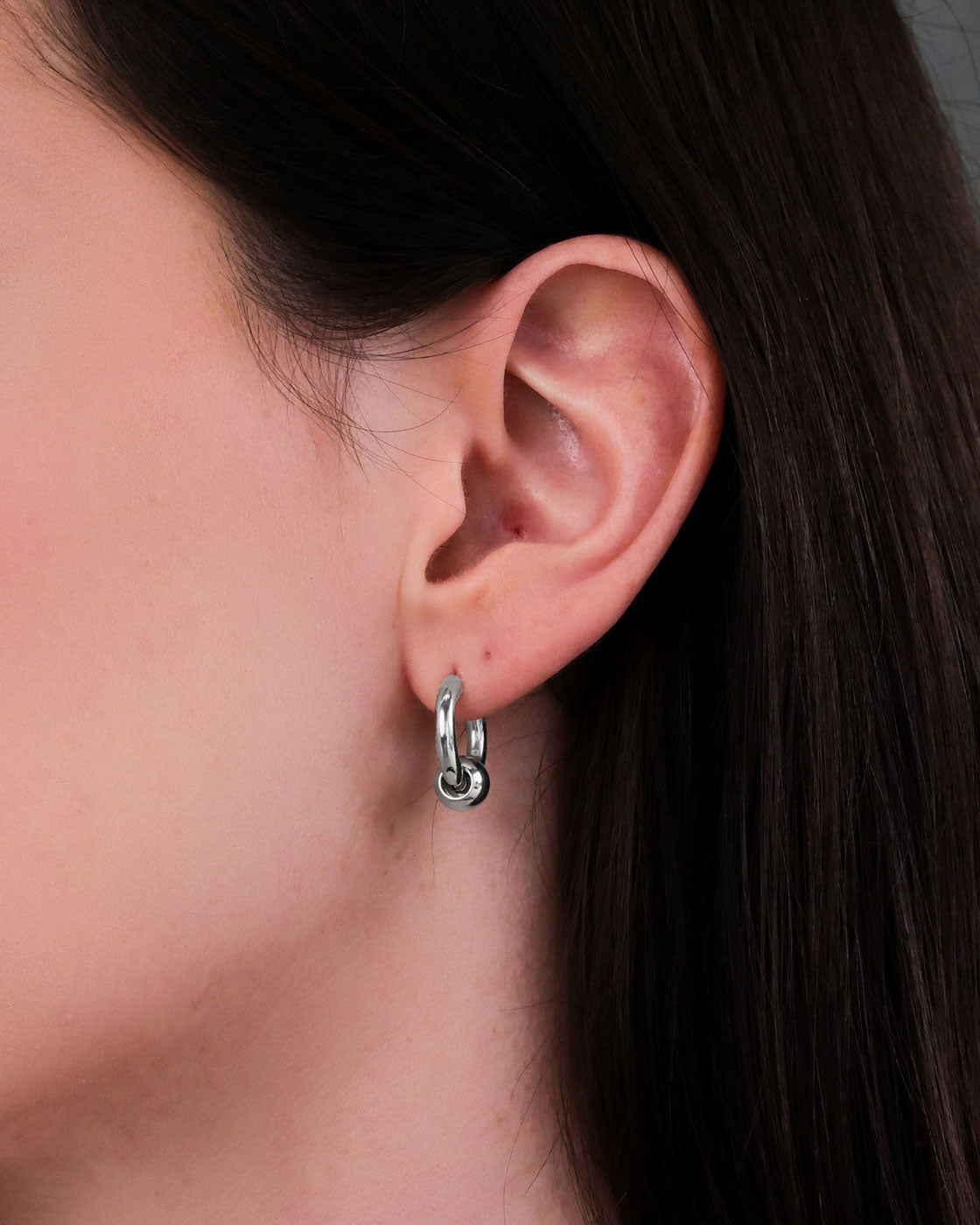 Close-up of a woman's ear with straight brown hair, wearing small hypoallergenic stainless steel Ball Hoop Earrings. The hoops feature a stylish knot detail, and the background is neutral and unobtrusive.