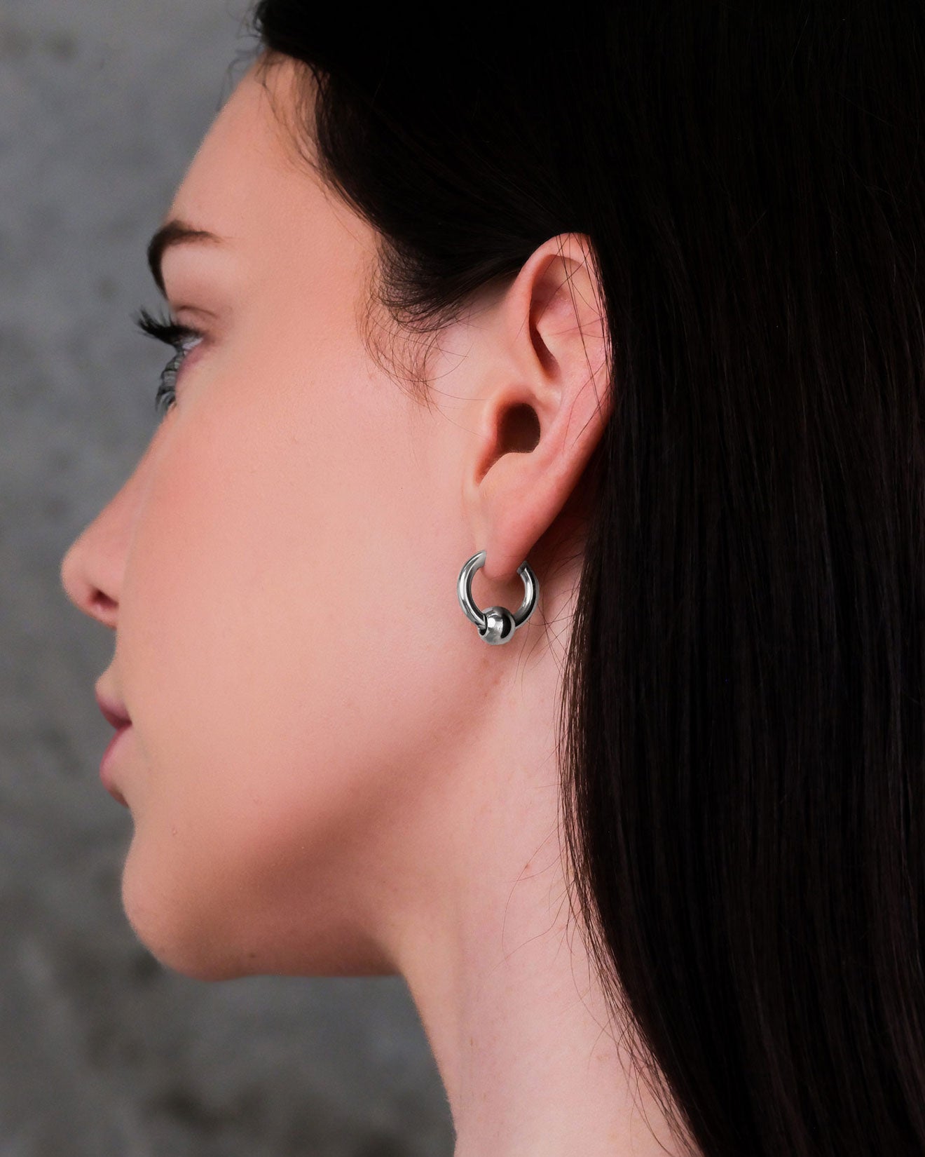 A close-up side view shows a woman with straight dark hair wearing Ball Hoop Earrings made of hypoallergenic stainless steel, highlighting the small hoop on her ear against a blurred gray background.