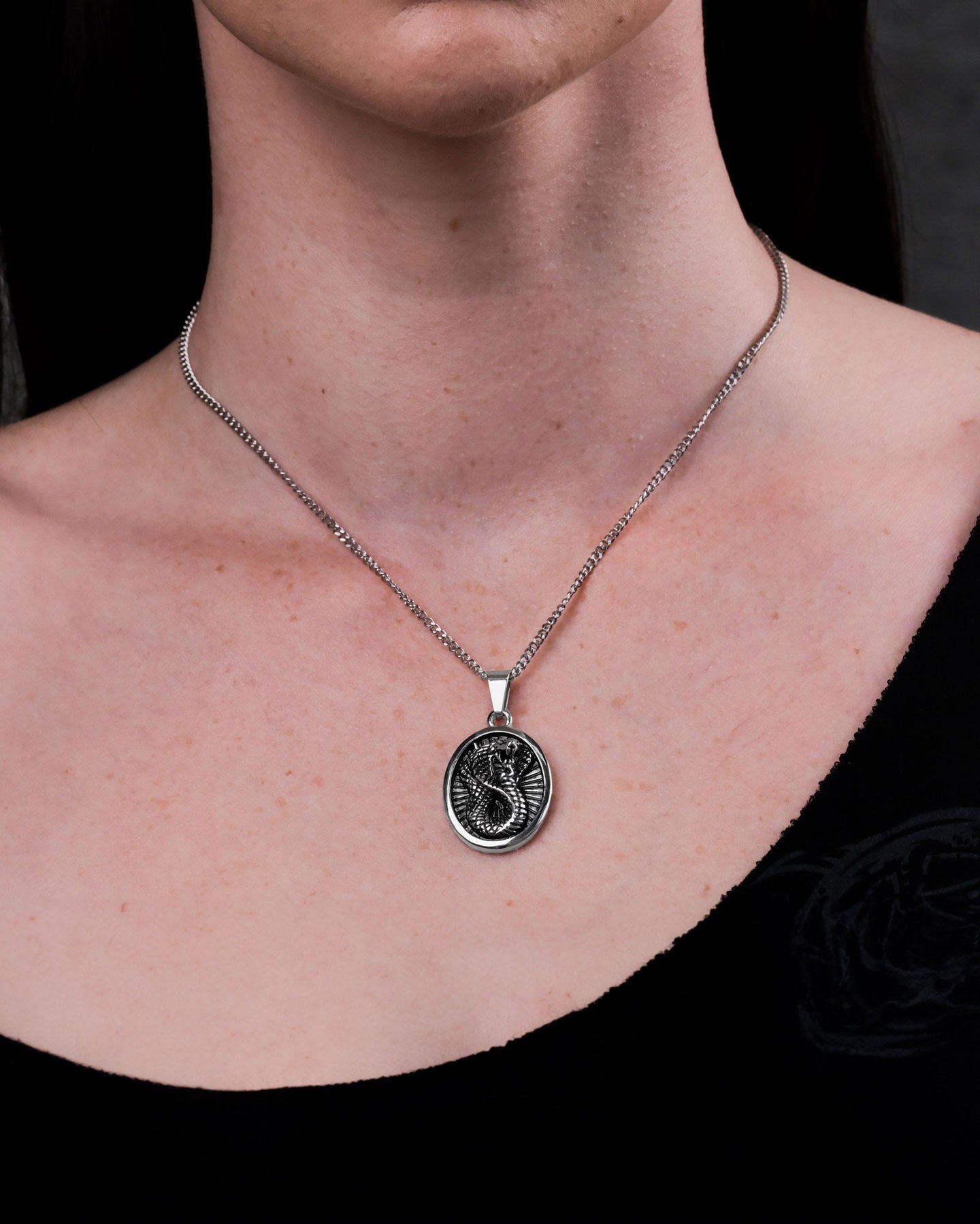 A person wearing the Snake Charmer Pendant—a silver chain necklace with a stainless steel oval pendant engraved with a dragon—photographed against a plain dark background.