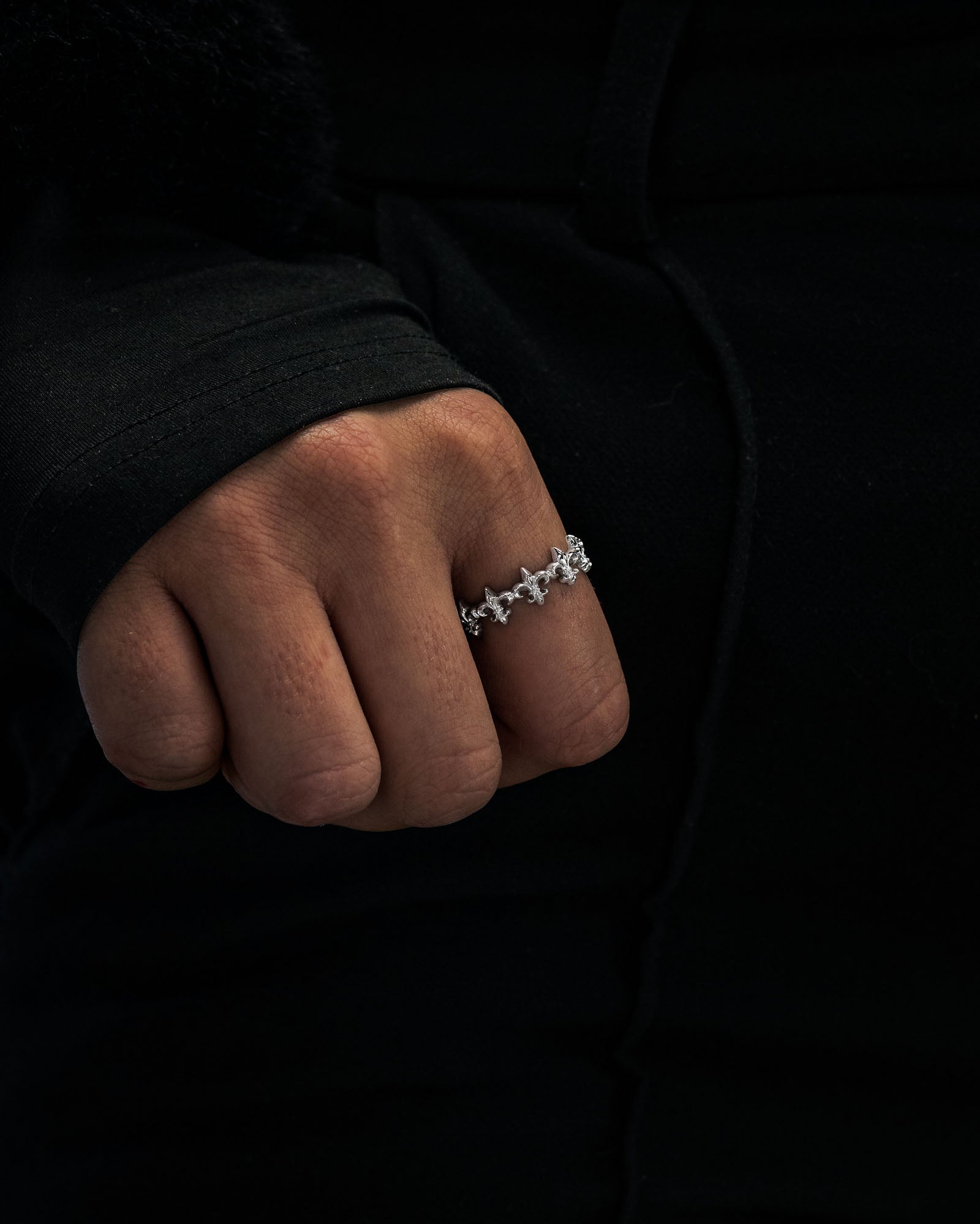 A hand adorned with Statement Collective’s “Fleur De Lis” ring, featuring a silver floral pattern, rests against a dark, long-sleeved garment.