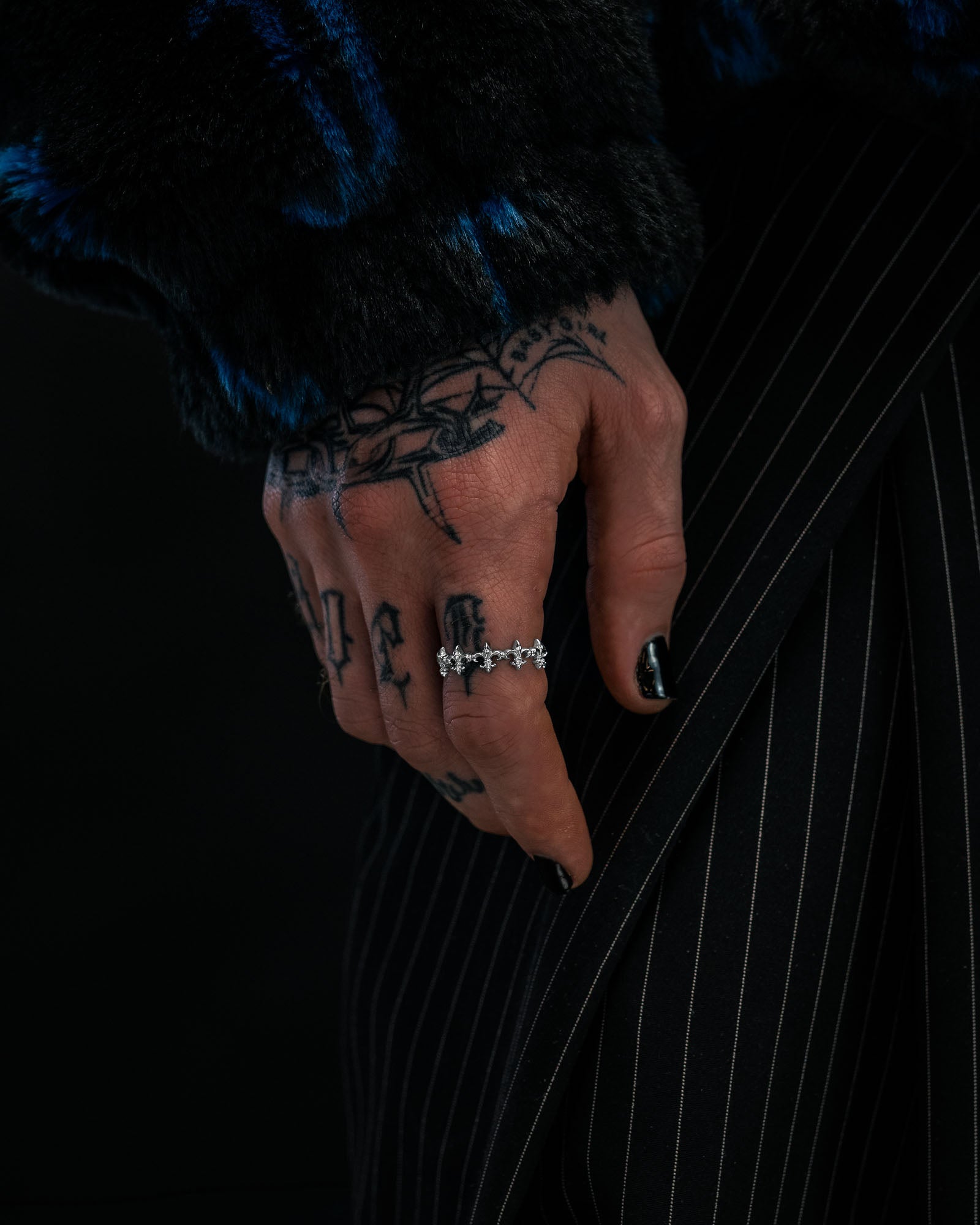 A tattooed hand with black nail polish wears the “Fleur De Lis” Ring by Statement Collective, a silver, stackable piece featuring star-shaped details, resting on pinstriped pants. The person also wears a dark, furry textured jacket.