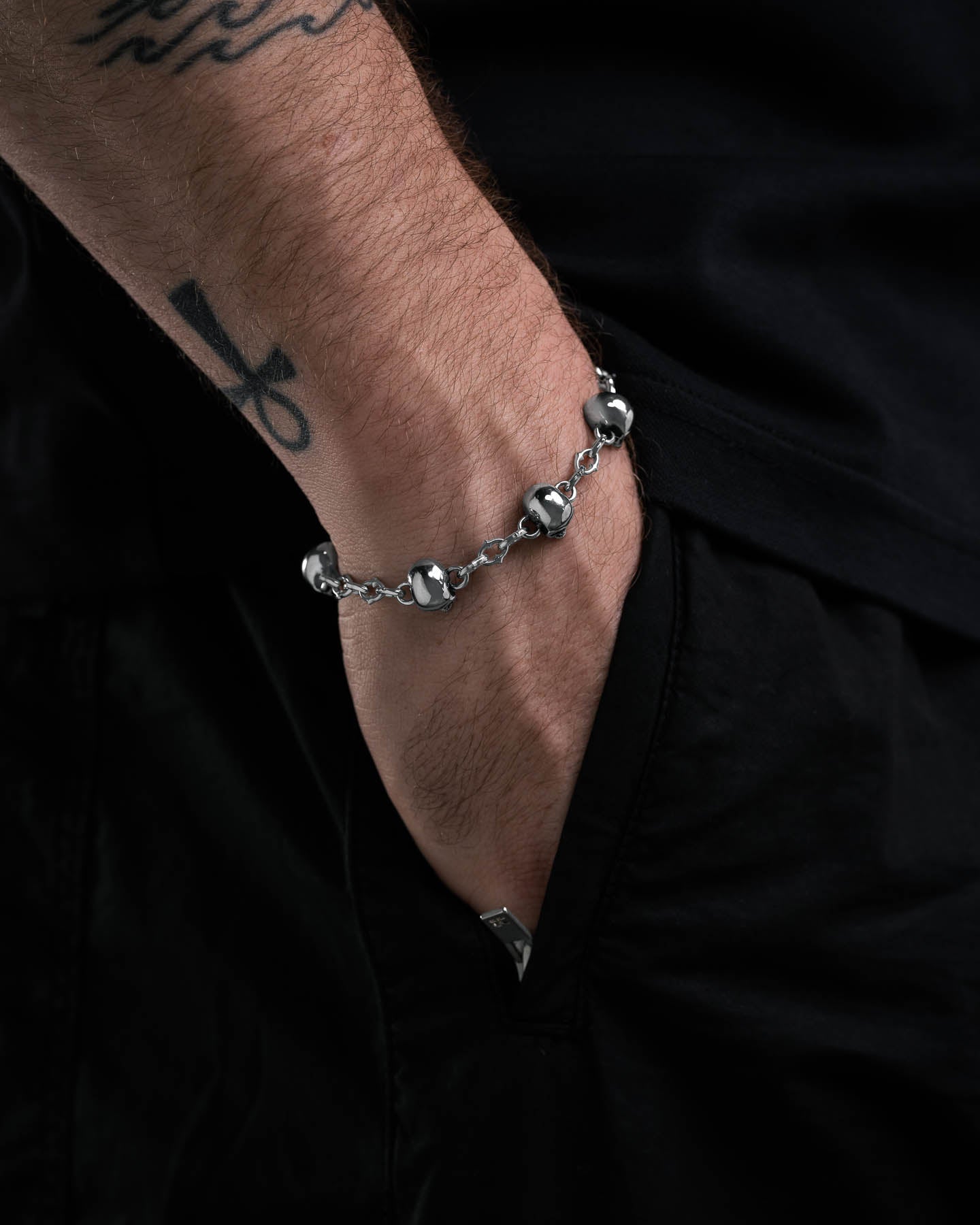A person with an ankh tattoo on their wrist wears the Skull Charm "Cathedral" Spiked Link Bracelet, showcasing their alternative style with their hand in the pocket of dark pants against a shadowy background.