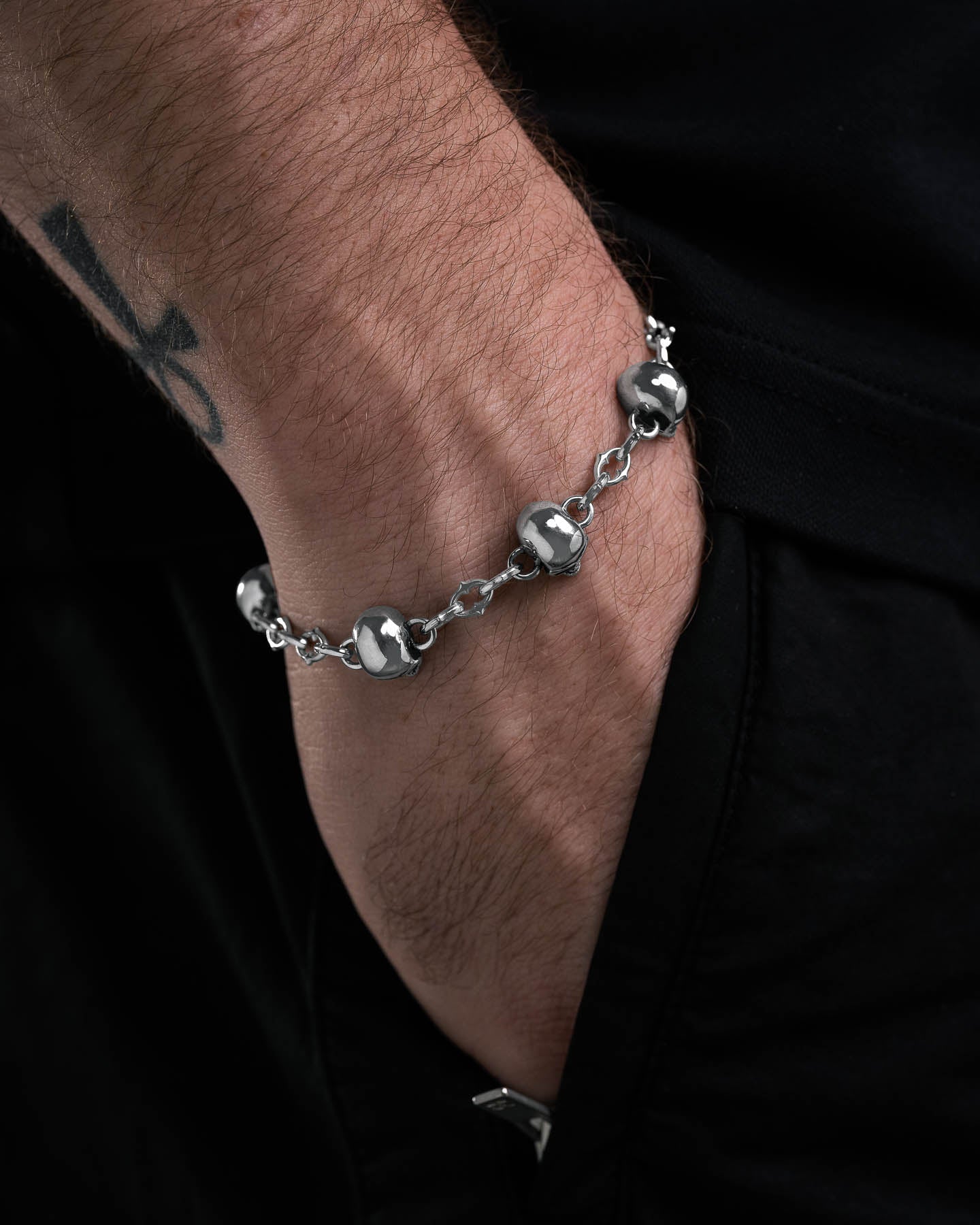 A close-up of a tattooed hand wearing the Skull Charm "Cathedral" Spiked Link Bracelet, partly tucked into black pants for an edgy alternative style.