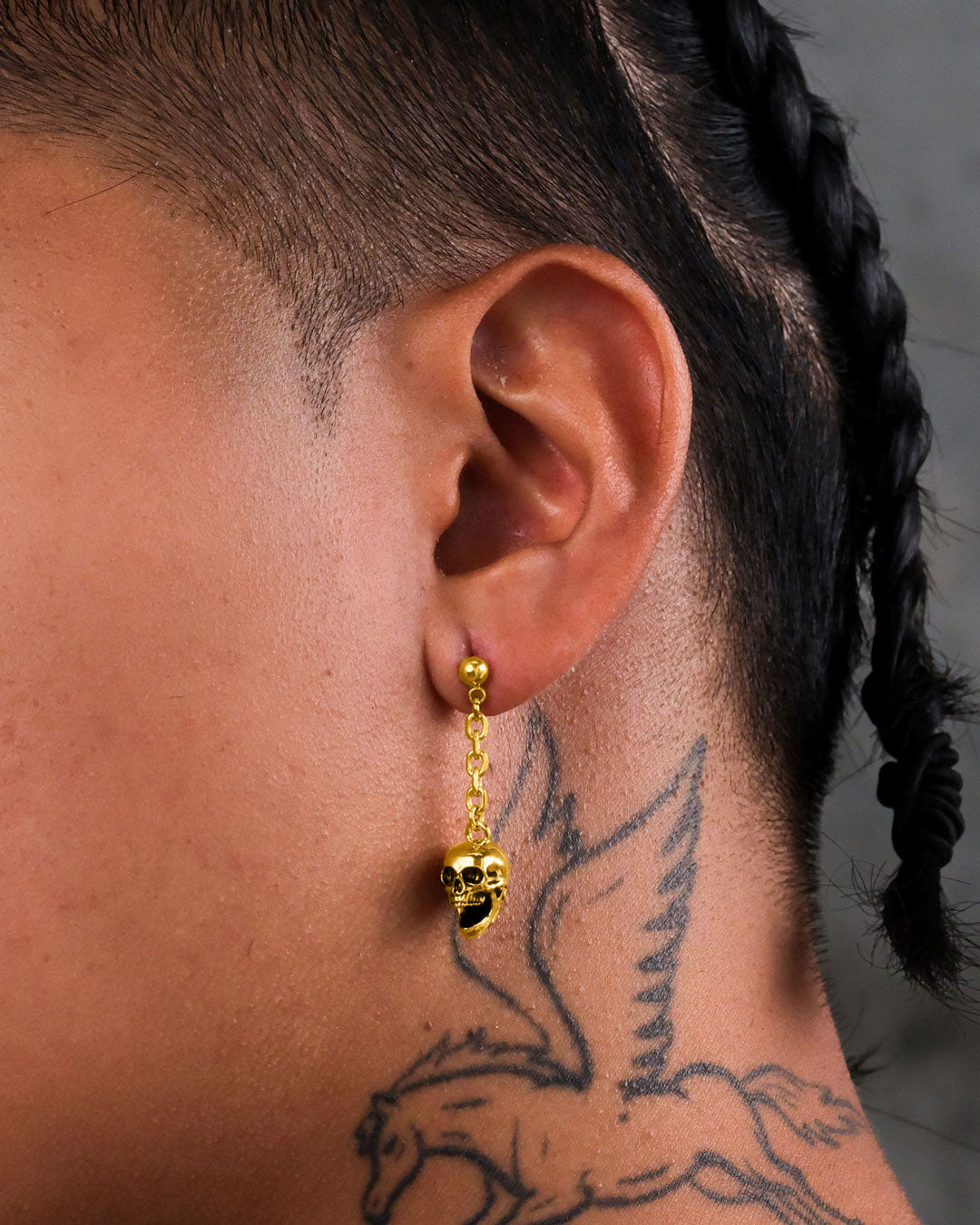 A close-up of an ear wearing hypoallergenic Skull Drop Earrings (Gold). The earring dangles elegantly; the person has braided hair and a winged horse tattoo on their neck.