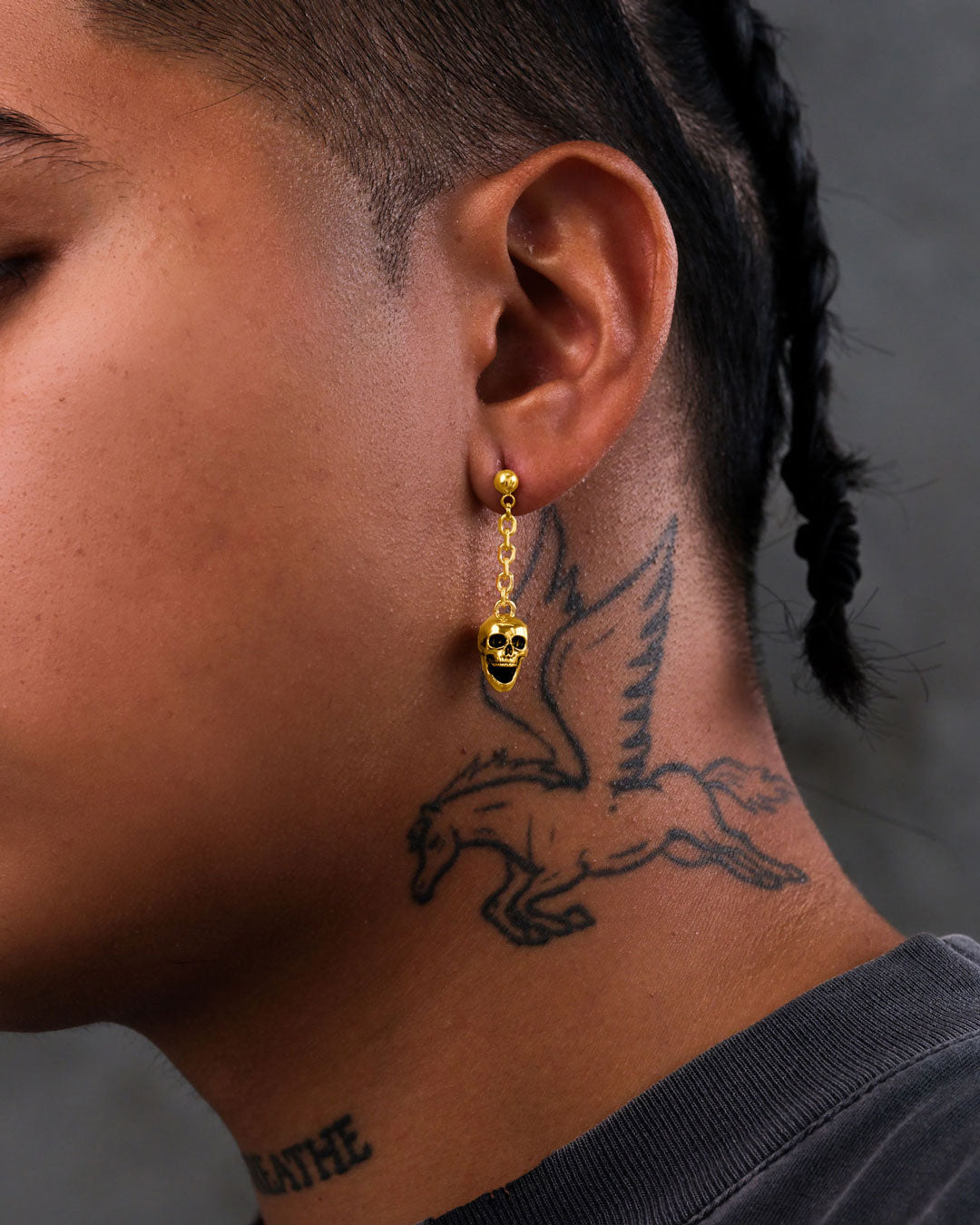 Close-up of a person’s ear wearing Skull Drop Earrings (Gold). They have a winged horse tattoo on their neck and braided hair. The background is plain dark gray.