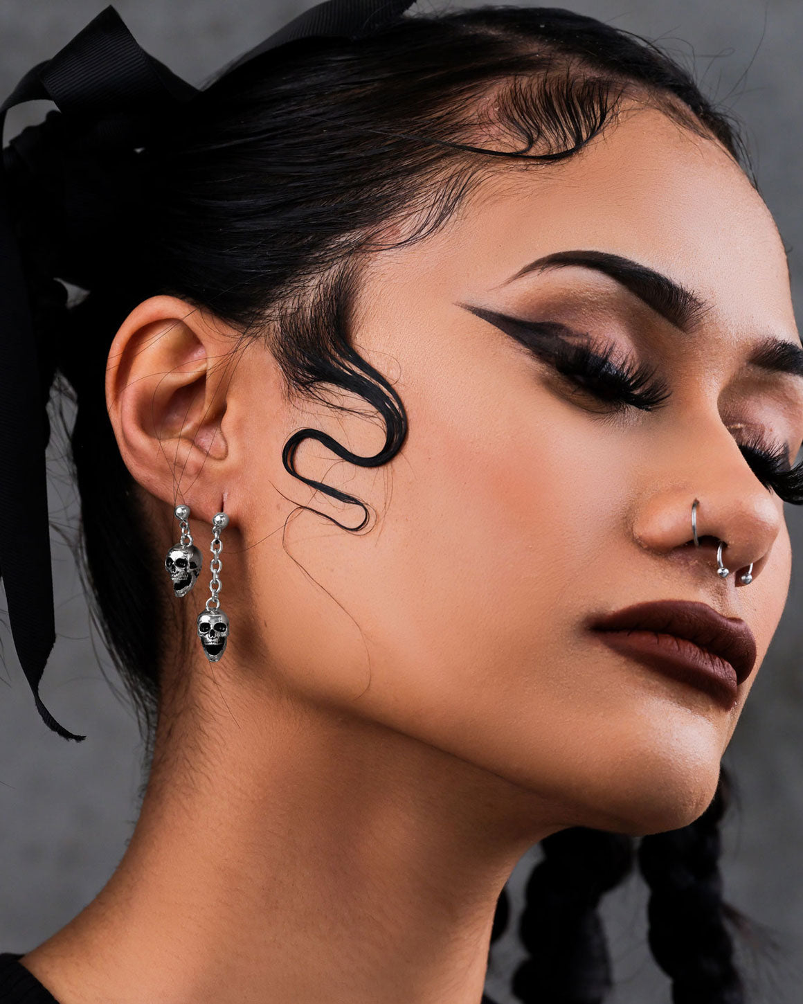 Woman with dramatic black winged eyeliner, dark lipstick, and styled baby hairs wears Skull Drop Earrings and a septum ring, eyes closed against a neutral background.