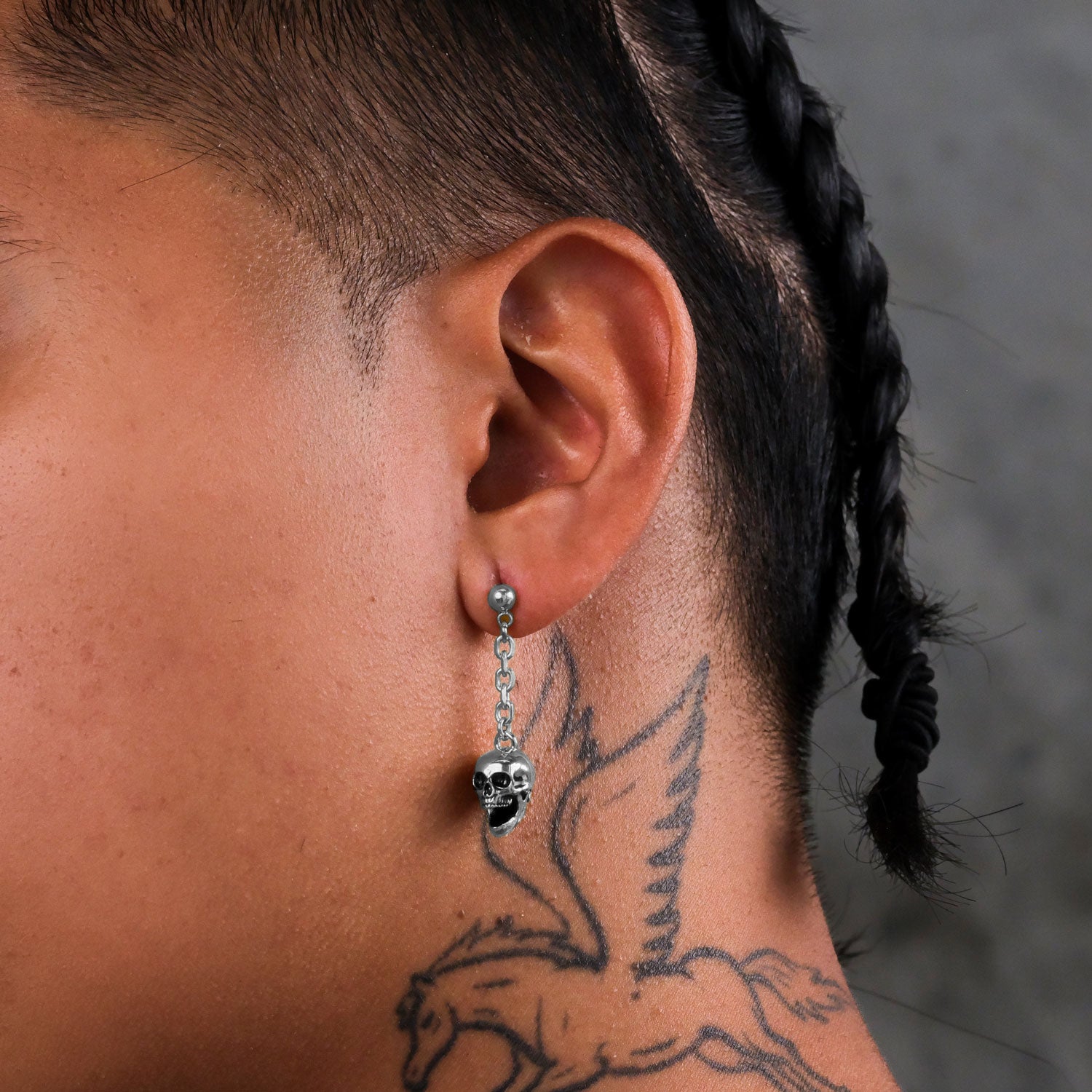 Close-up of a person with braided hair and a winged horse neck tattoo wearing Skull Drop Earrings. Made from hypoallergenic stainless steel, the dainty drop earrings are showcased against a blurred background.
