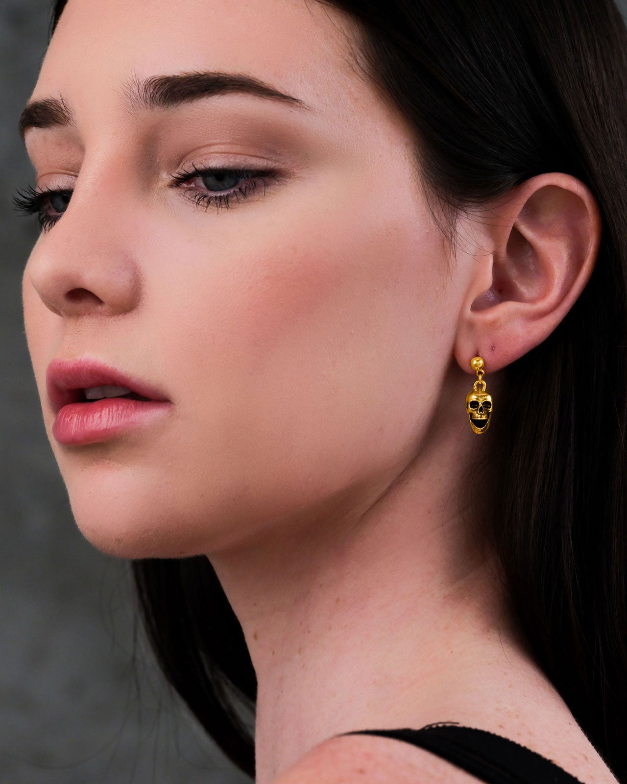 A woman with fair skin and dark hair wears minimal makeup and Skull Earrings (Gold). The 18k gold-plated punk jewelry pops against her look, while the blurred gray background keeps the focus on her standout accessories.