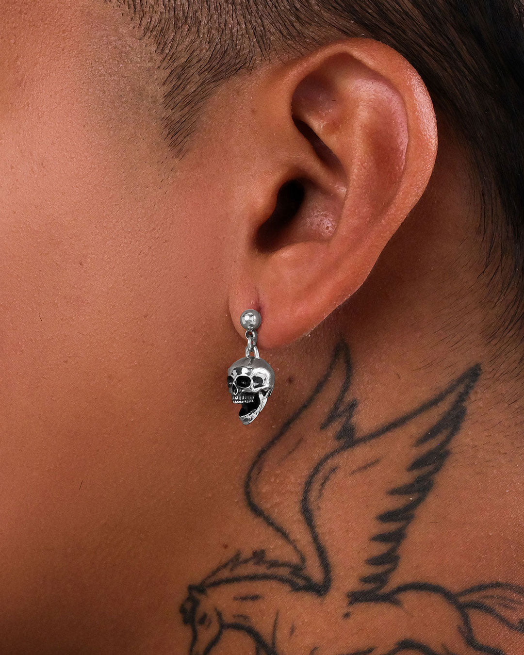 A close-up of a person's ear wearing Skull Earrings, showcasing rock fashion jewelry style, with a flying bird tattoo visible on their neck.