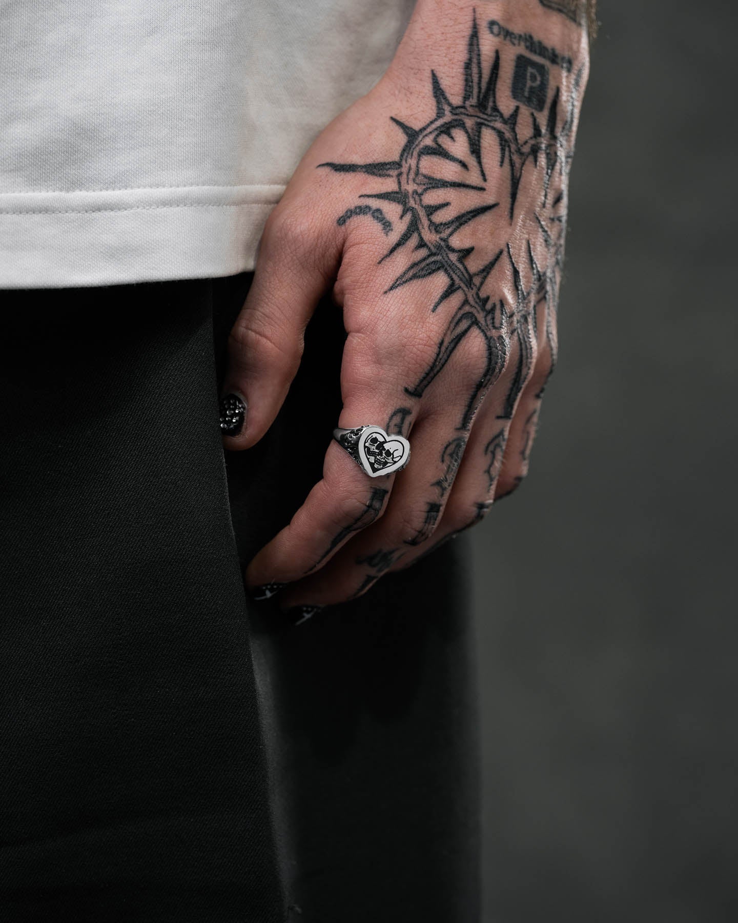 A close-up of a tattooed hand by their side shows a large heart with rays tattoo, black nail polish, and the Skull Heart Ring—an edgy gothic jewelry piece. The person wears dark pants and a light shirt.