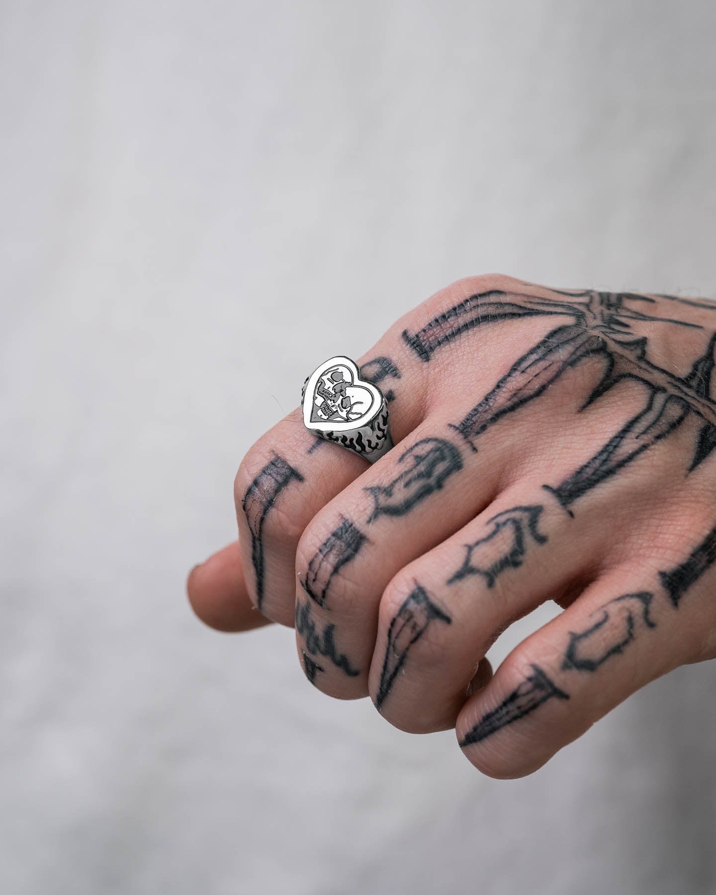 A hand with skeleton tattoos wears the Skull Heart Ring—silver, heart-shaped gothic jewelry with a black illustration—set against a plain, light background.