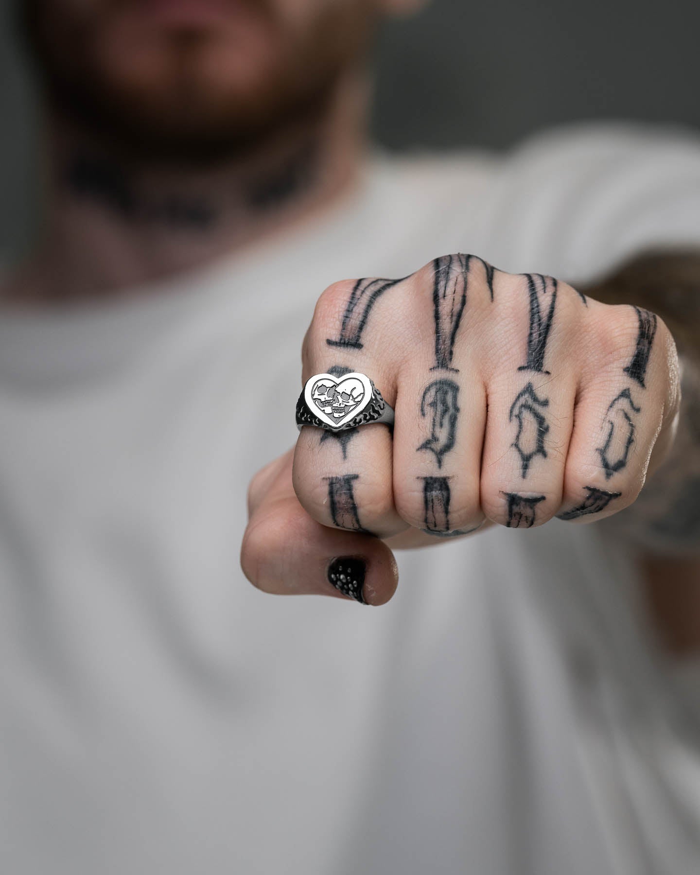 A person in a white shirt holds a fist forward, showcasing the Skull Heart Ring on their finger. The image focuses on the hand and ring, with black arrow tattoos visible and the face blurred, highlighting this unique gothic jewelry piece.