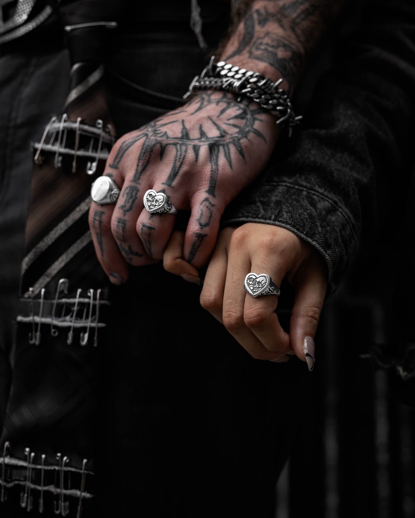 Two hands, one tattooed and both wearing gothic jewelry—silver rings with heart motifs. The tattooed hand features the Skull Heart Ring, a bracelet, edgy safety pin and chain-accented clothing; the other has manicured nails.
