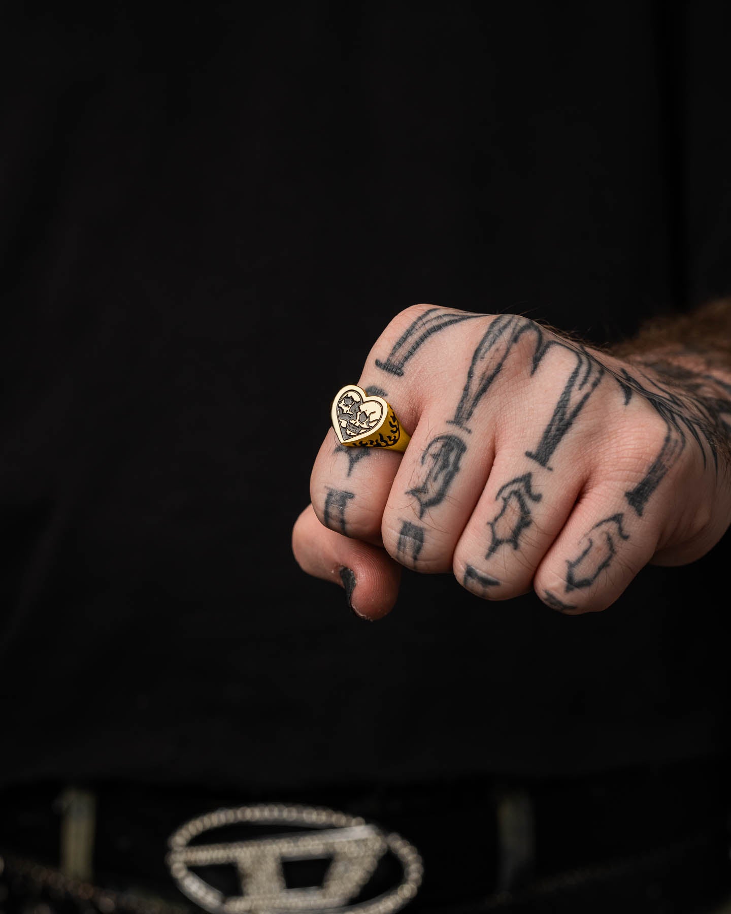 A close-up of a tattooed hand wearing the Skull Heart Ring (Gold) with 18k gold plating. The person, dressed in black, shows part of a decorative belt buckle, completing the bold gothic jewelry look.