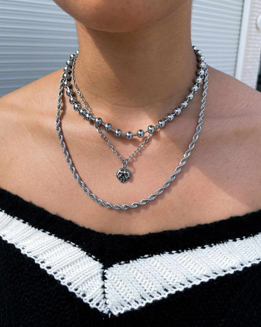 A person wears three layered silver necklaces—a round bead chain, a twisted rope chain, and the Skull Heart Pendant—paired with a black and white off-shoulder top for a dark romance jewelry look.