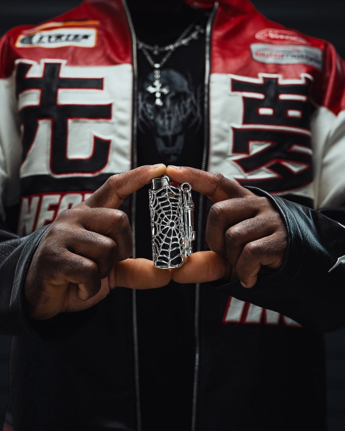 A person wearing a red, white, and black jacket holds the Statement Collective Spiderweb Lighter Sleeve in silver between their hands, with part of their necklace and shirt visible in the background.