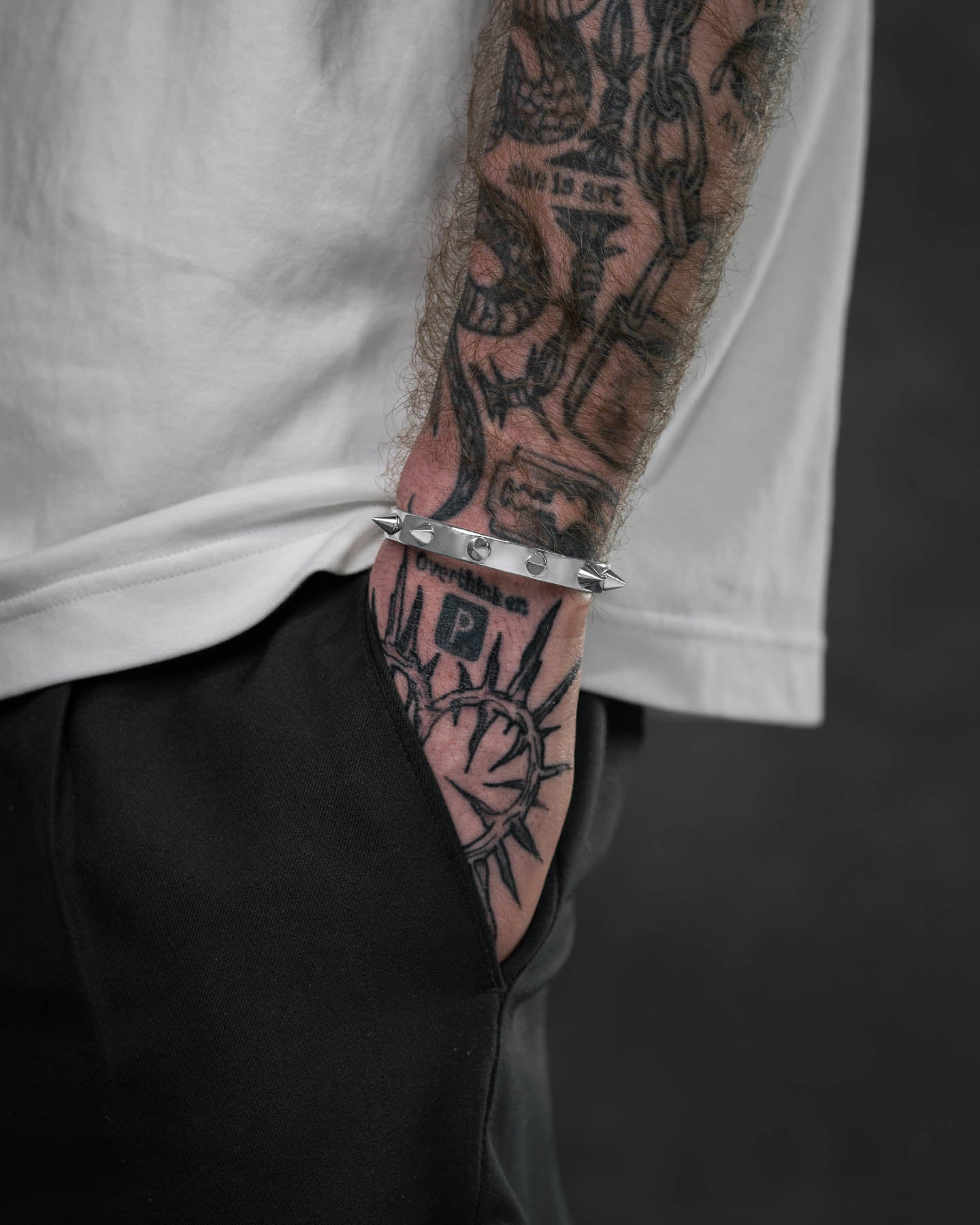 Wearing a white shirt and black pants, a person shows off a tattooed forearm and hand adorned with the Spiked Cuff Bracelet. The bold tattoos and the edgy bracelet create a striking, statement look.