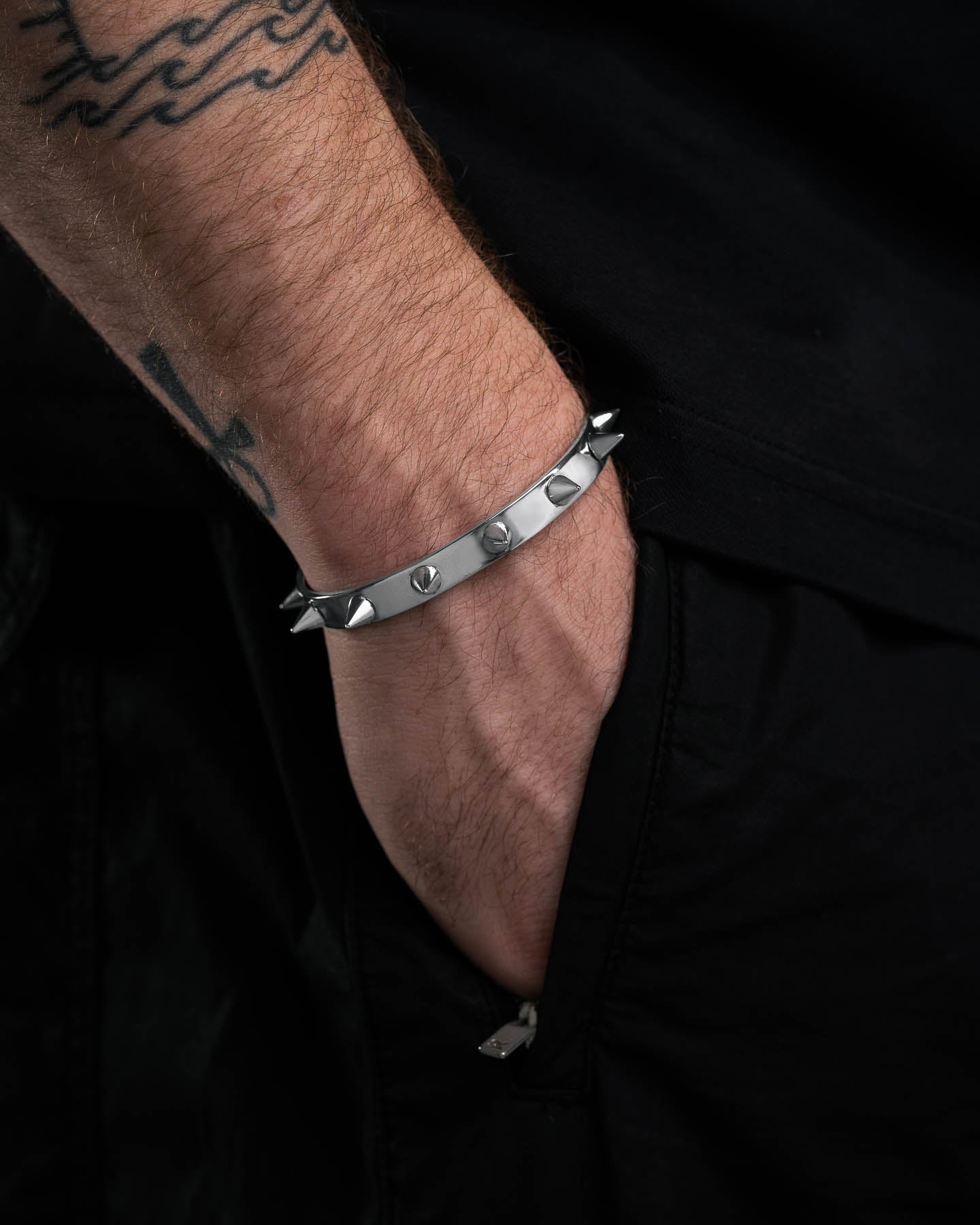 A close-up shows a tattooed forearm and hand in a black pocket, with the bold Spiked Cuff Bracelet standing out against all-black clothing.