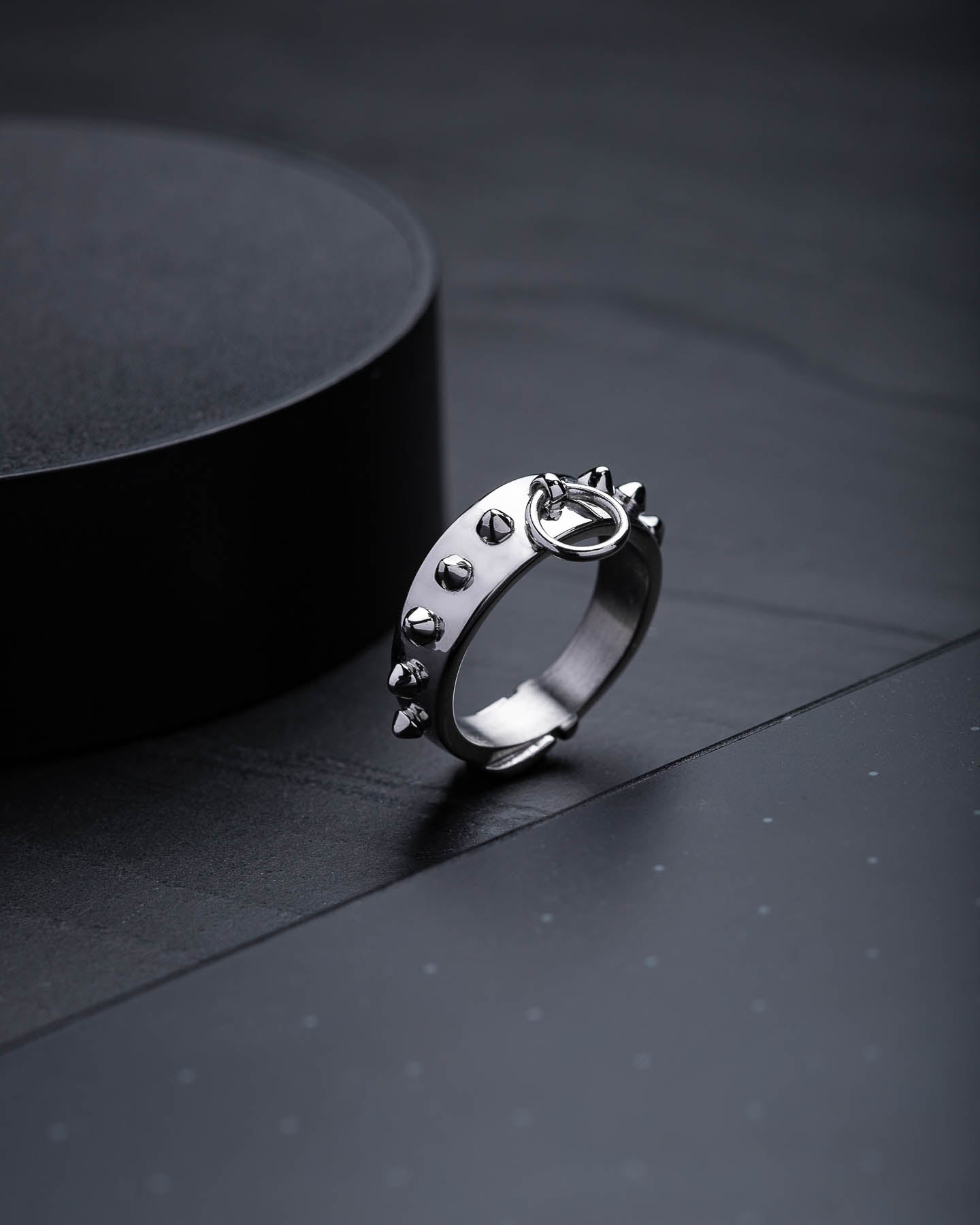 The Spiked Dog Collar Ring, crafted from stainless steel with stud accents and a bold circular loop detail, stands upright on a dark textured surface with a black cylinder behind it, adding an edgy vibe to goth jewelry collections.