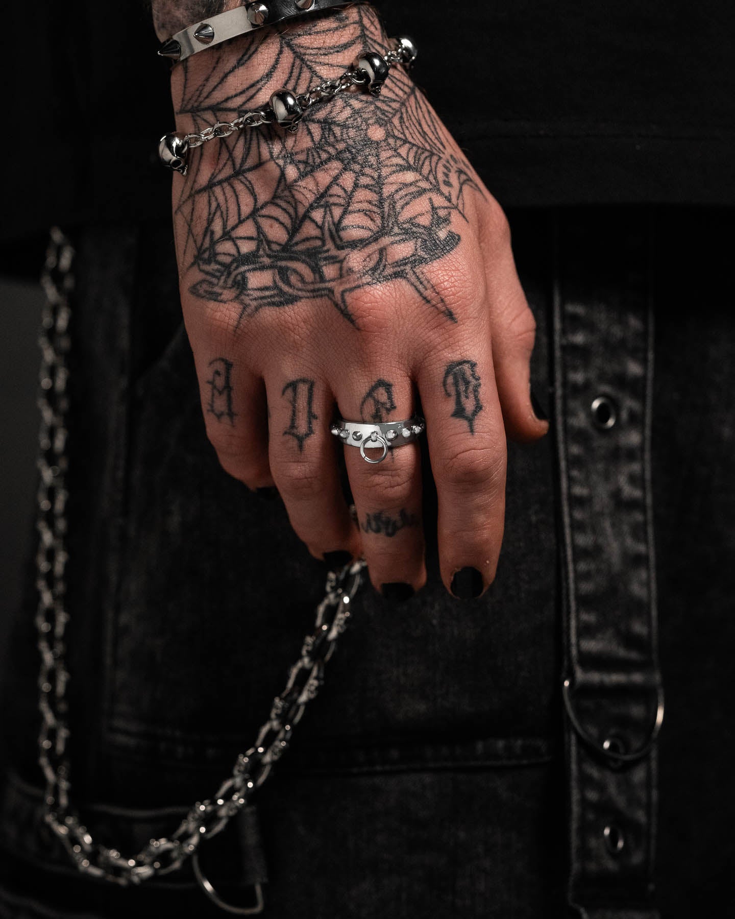 A close-up of a tattooed hand with bold letter and spiderweb tattoos, silver rings, black nail polish, and goth jewelry including the Spiked Dog Collar Ring, resting on black denim pants with chains.