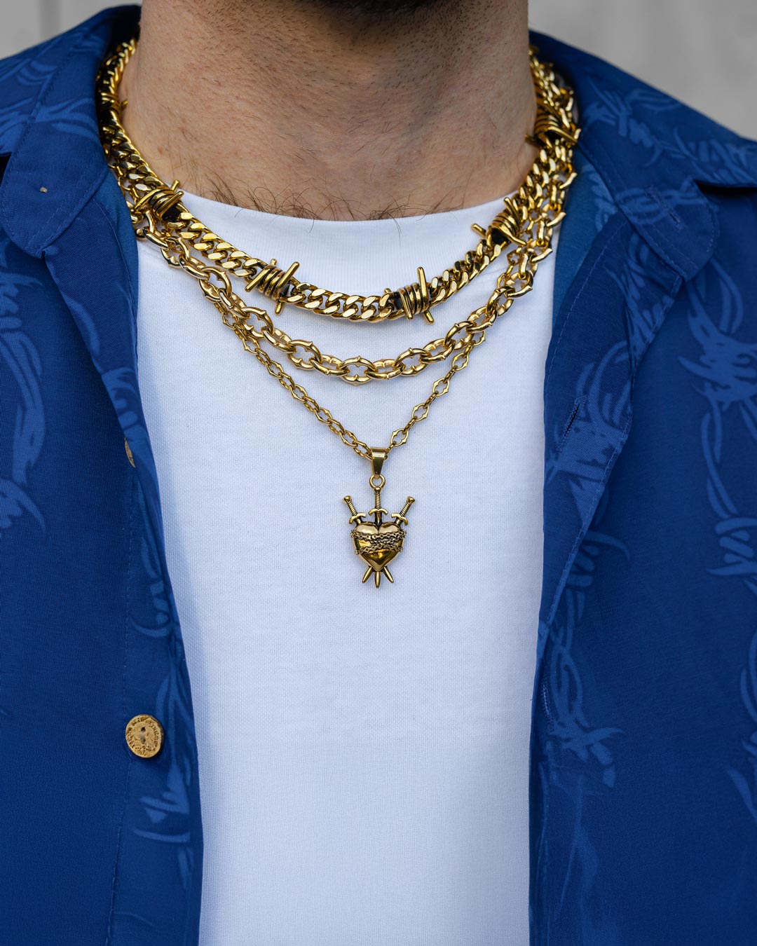 A person wears three gold chains, including the Spite Stack Gold (Custom Pendant Choice) necklace, layered over a white T-shirt and blue shirt. The photo is cropped to focus on the neck and upper chest.