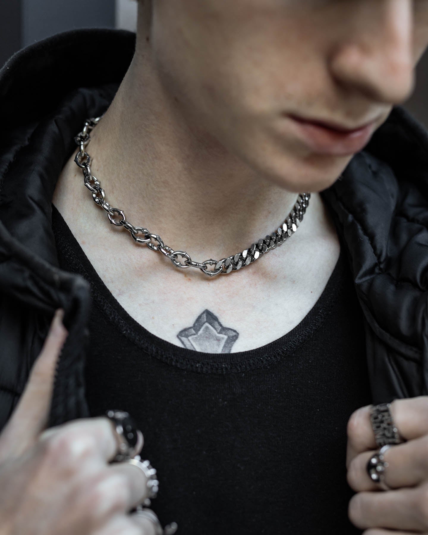 A person wearing the "Split" Cathedral Cuban Chain (Adjustable) with a black shirt and jacket. They have a star tattoo on their upper chest, several rings for a gothic vibe, and their face is partially out of frame.