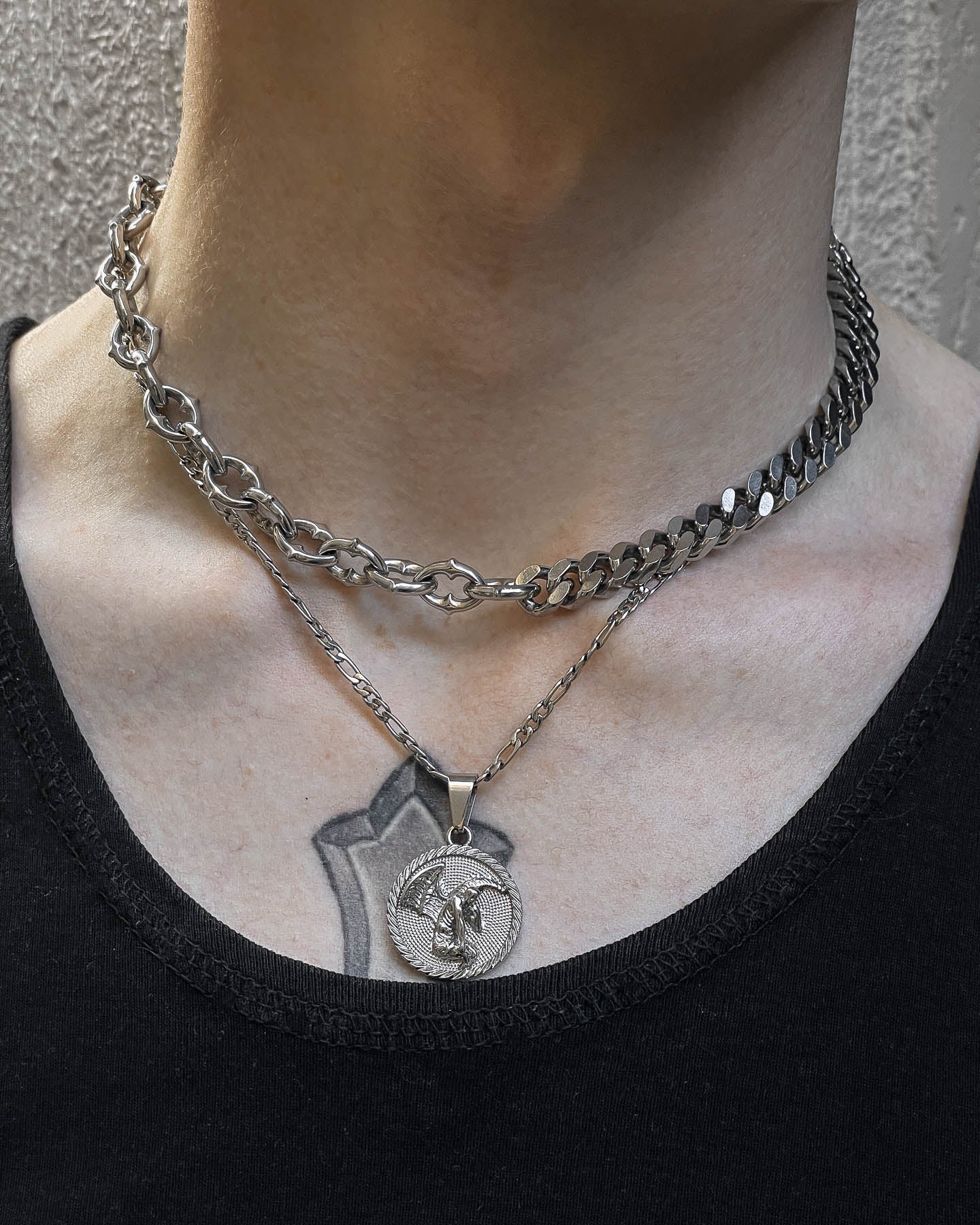 A person wears the "Split" Cathedral Cuban Chain (Adjustable), a chunky silver necklace, layered with a thinner chain featuring a circular pendant. A tattoo peeks from their upper chest above their black top.
