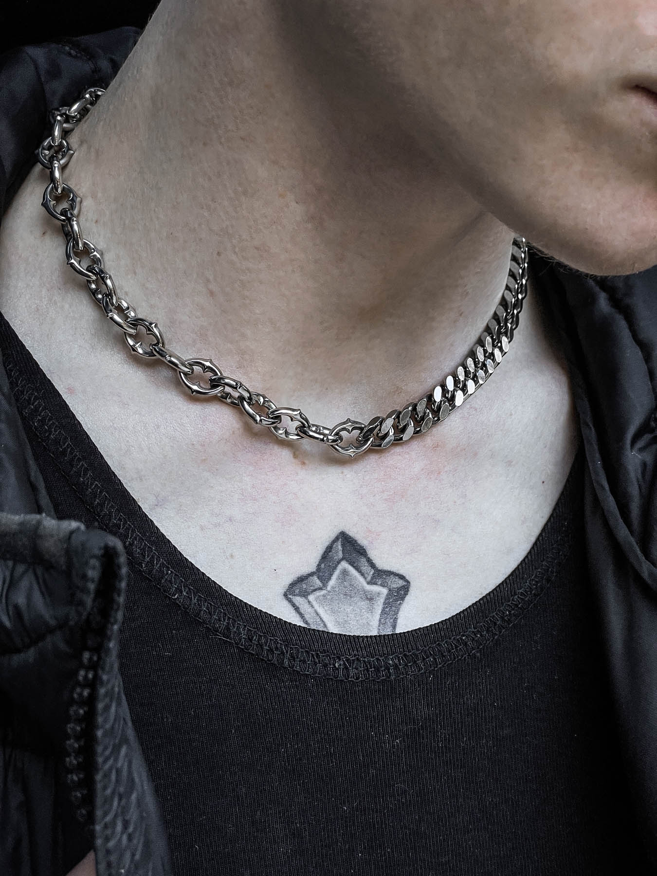 A close-up of a person wearing the "Split" Cathedral Cuban Chain (Adjustable) with a black shirt and dark jacket; a geometric star tattoo is visible on their upper chest near the necklace's neckline.
