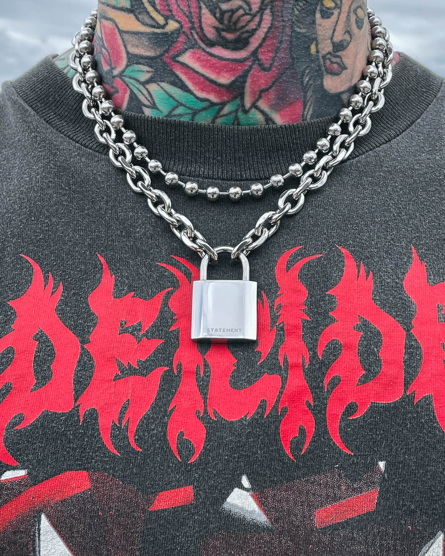 Wearing Statement Lock & Chain 2.0, a person with colorful neck tattoos layers two chunky silver necklaces with a bold padlock pendant. Their black shirt, decorated with vivid red flames and stylized text, highlights the striking jewelry.