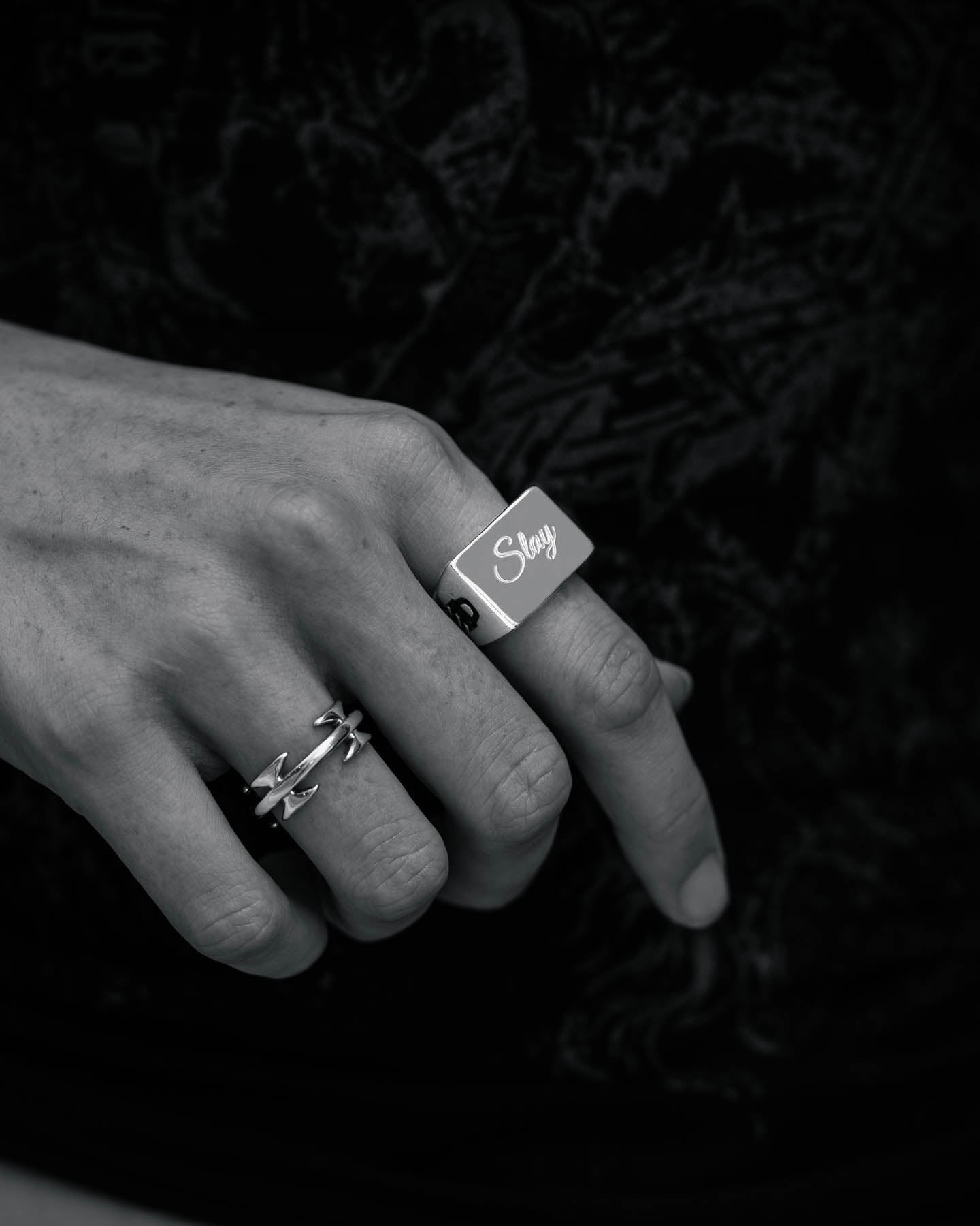 A close-up black-and-white photo shows a hand wearing two rings: the Statement Signet Ring, a chunky stainless steel piece engraved with "Slay," and a thinner crisscross ring, both set against a dark, textured background.