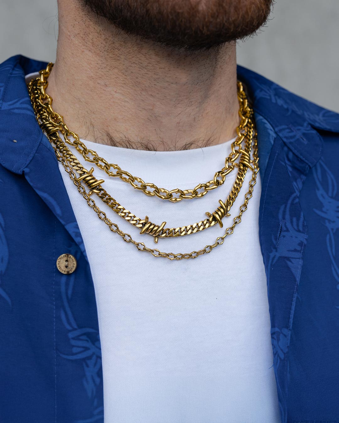 A person in a white shirt and blue button-up is shown wearing the Stay Safe Stack (Gold) necklace, featuring three layered gold chains with spiked and barbed wire-style details. The image focuses on the neck and upper chest.