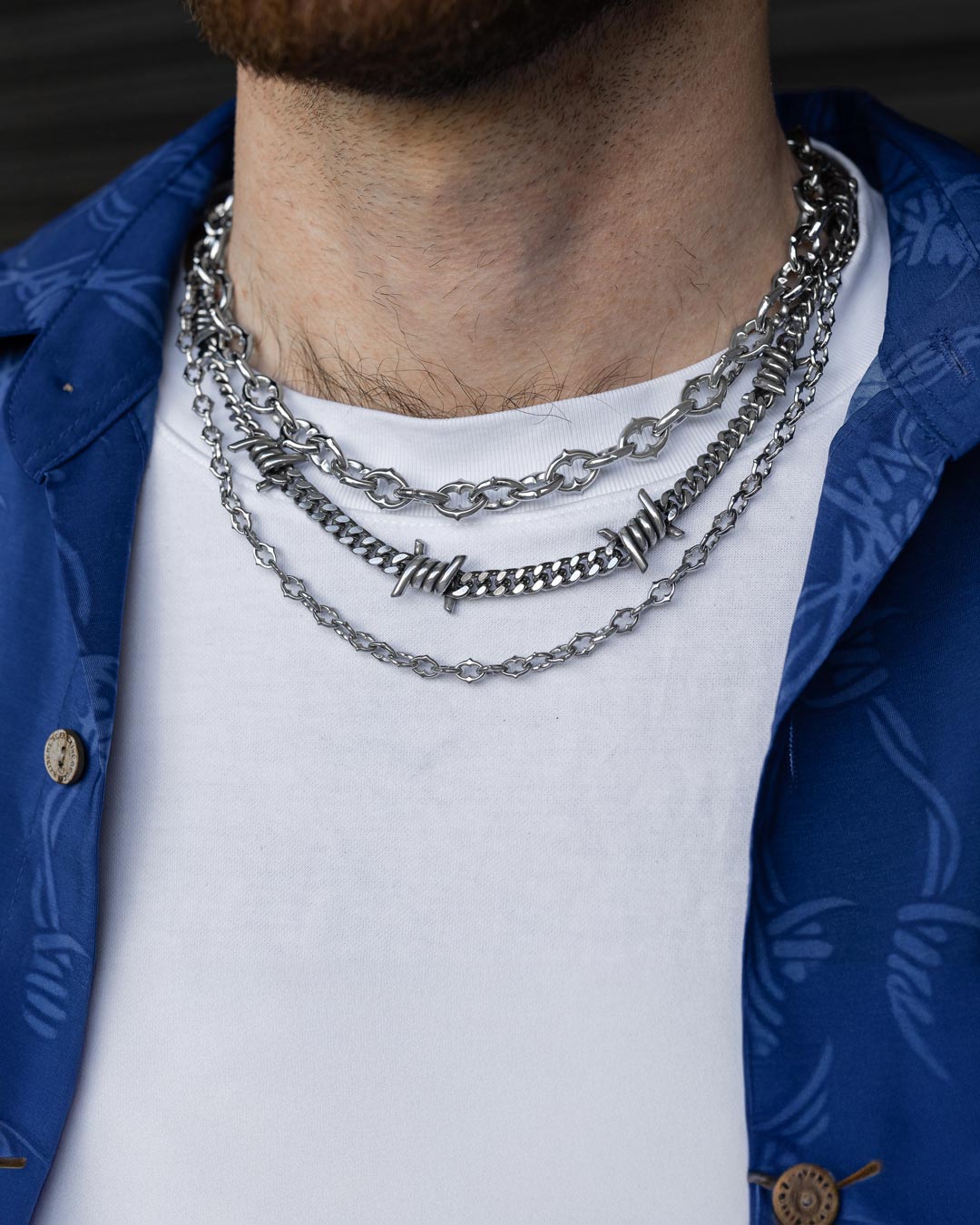 A close-up of a person wearing the Stay Safe Stack—a bold mix of layered silver chains, spiked chains, and a barbed wire necklace—styled over a white t-shirt and blue patterned open shirt.