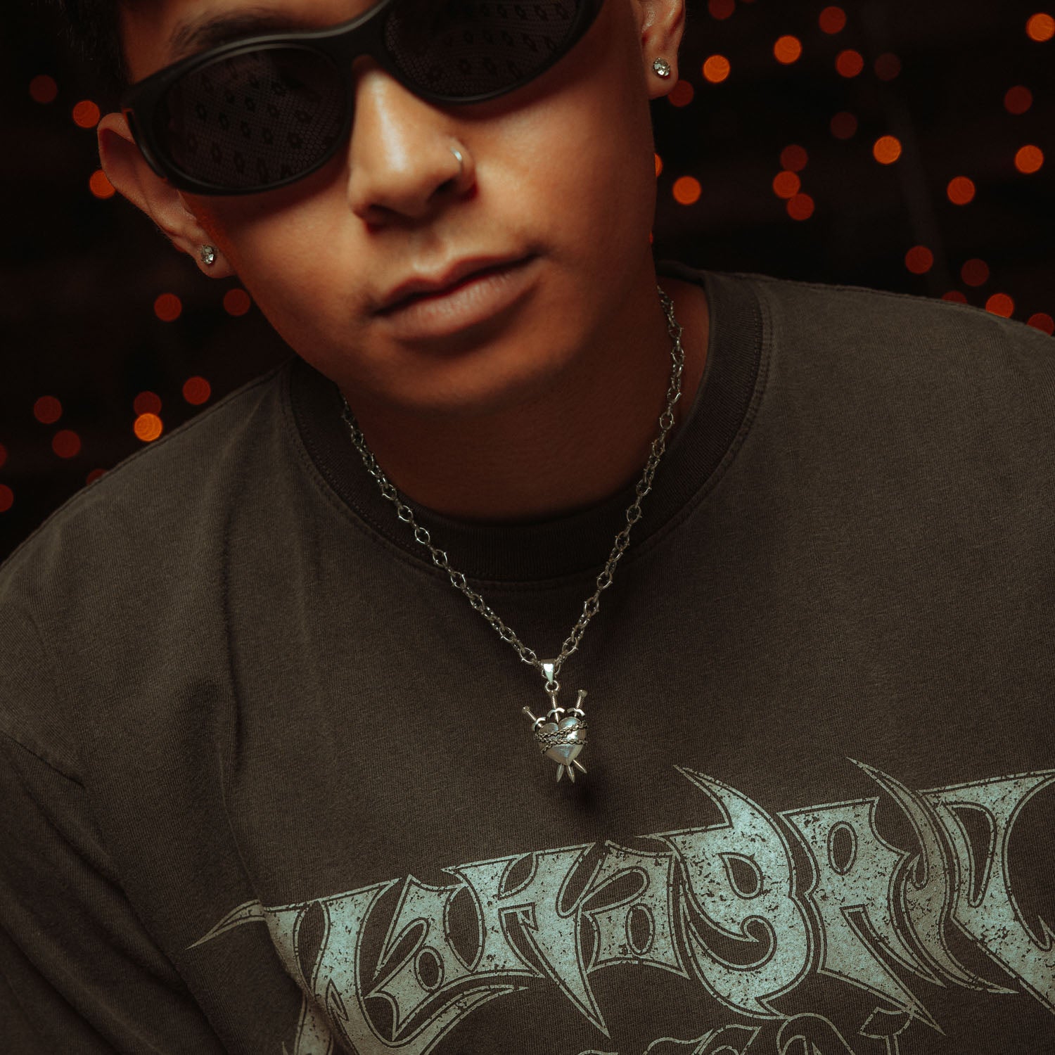 Silver heart pendant with three swords going through it and chain wrapped around it on a spiked chain on male model in gret shirt and black sunglasses